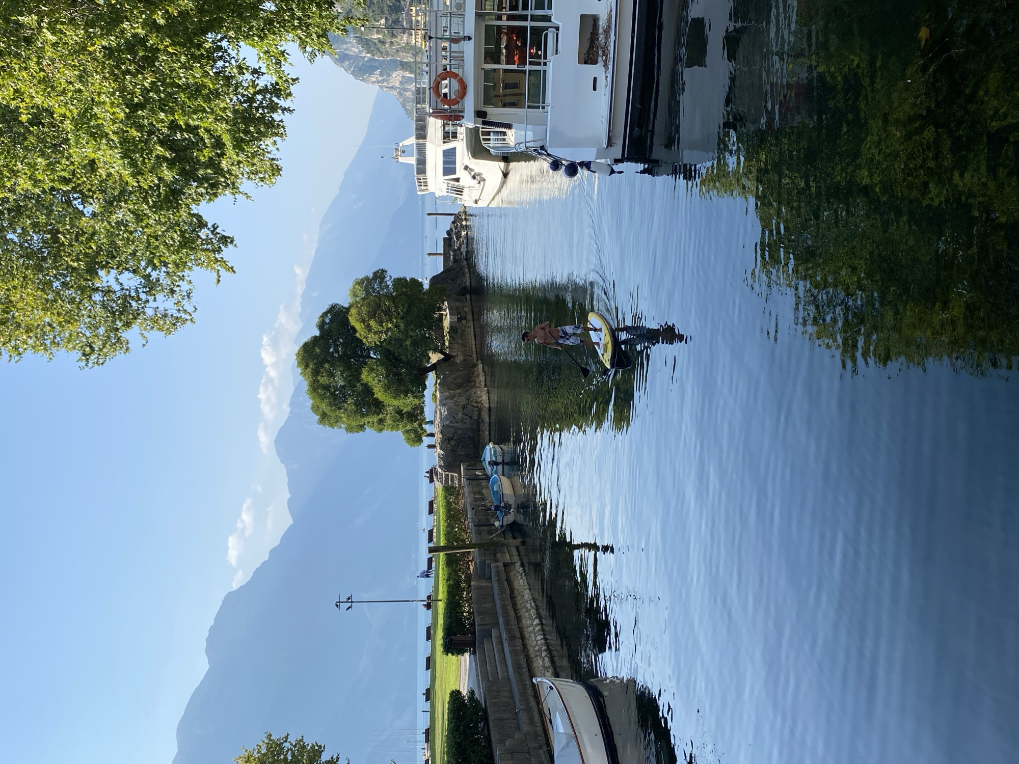 The image captures a serene scene at the harbor of Riva del Garda, Italy. In the foreground, the calm, blue water reflects the clear sky and surrounding greenery. A person is seen paddleboarding in the middle of the canal, standing on a yellow board and holding a paddle. To the right, two white boats are docked, one larger with multiple decks and a life preserver visible, and another slightly smaller. On the left bank, a stone wall lines the canal, with steps leading up to a grassy area. Small boats are tied to a wooden post along the wall. In the background, a line of benches overlooks a tranquil lake, with a distant view of majestic mountains under a bright blue sky. Lush green trees frame the top right of the image, adding to the natural beauty of the location.