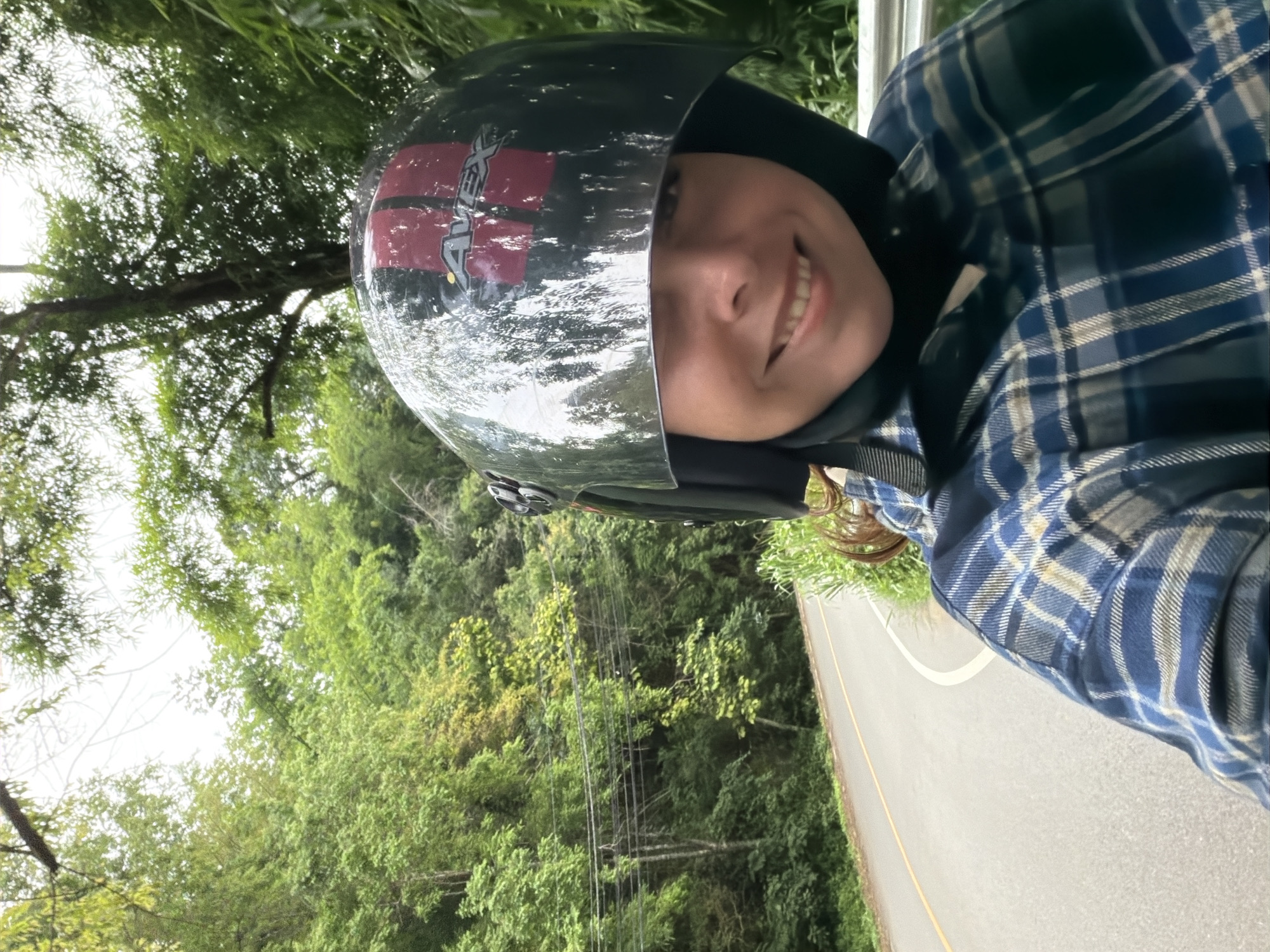 A person is on a motorcycle on a road in Samoeng Tai, Thailand. The person is wearing a helmet and a blue plaid shirt. The road is paved and has a yellow line down the middle. The surrounding area is a lush, green forest with tall trees. The sky is overcast.