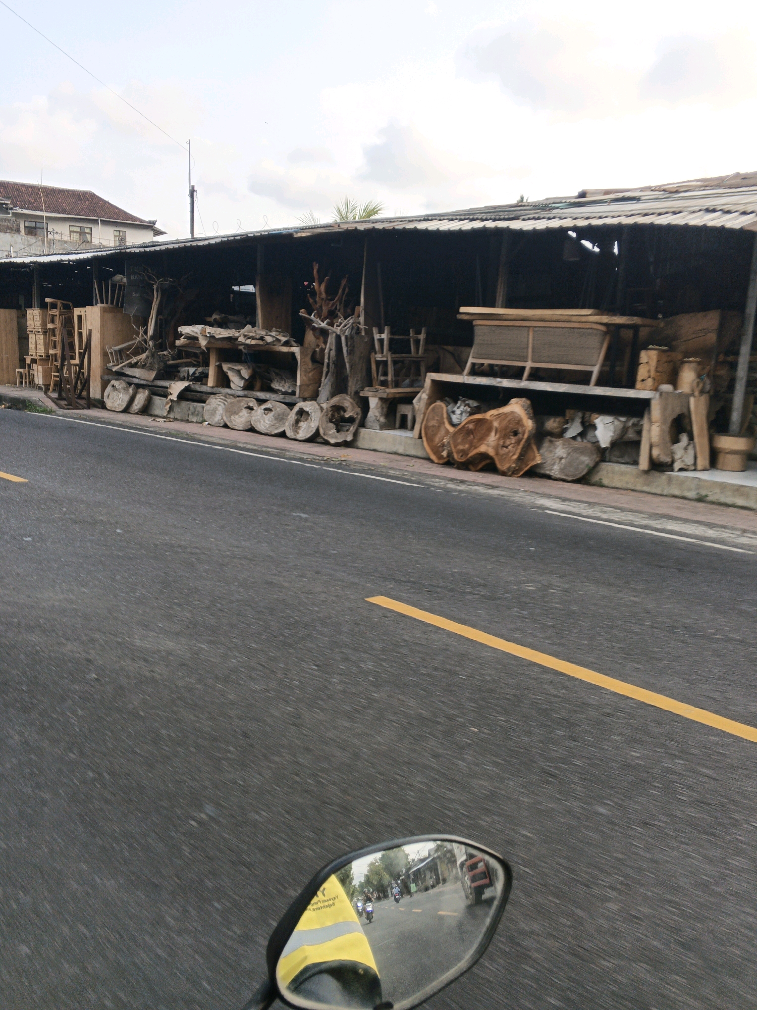 The image depicts a roadside scene in Sukawati, Indonesia, likely a street in the artistic region known for its wood carvings and furniture. In the foreground, a portion of a motorcycle's rearview mirror is visible, reflecting a street scene. The mirror shows a yellow and grey safety vest on the rider, and a reflection of other motorcycles and a building with shops. The road itself is asphalt with a visible yellow dividing line.
To the right of the road, a long, covered structure serves as a display area for various wooden furniture and raw wood pieces. The structure has a corrugated metal roof supported by wooden beams. Various unfinished and finished wooden items are arranged along the side of the road and under the shelter. These include large, circular wood slices, intricately carved wooden sculptures, tables, chairs, and benches. The wood displays suggest a workshop or a shop selling handcrafted wooden goods.
Behind the roadside display, a building with a red-tiled roof is partially visible, indicating a more permanent structure in the background. The sky above is partly cloudy, with a gentle haze. The overall impression is that of a bustling commercial area focused on woodworking crafts, characteristic of Sukawati's artistic heritage.