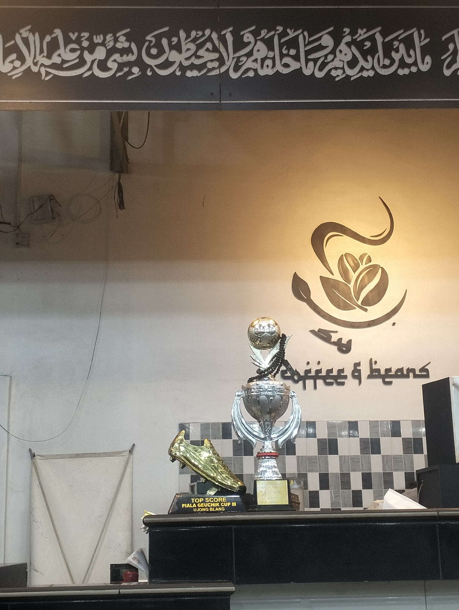 The image shows the interior of a coffee shop in Ujong Blang, Lhokseumawe, Indonesia. On a counter, there are two trophies, one with a golden soccer shoe and the other a silver cup topped with a soccer ball. The text on the plaque of the golden soccer shoe trophy reads "TOP SCORE PIALA GEUCHIK CUP III UJONG BLANG". Above the counter, on the wall, there is a coffee bean logo and text in Arabic, "كمعظ و عع coffee & beans". Above the logo and text, there is an inscription in Arabic calligraphic script, "ل ما بين ايدينَ مَا خَلَقَهُ وَ لَا يُحِيطُونَ بِشَى مَنْ عَلم الاعا". In the background, there is a white panel and a tiled wall. Electrical wires are visible on the wall.