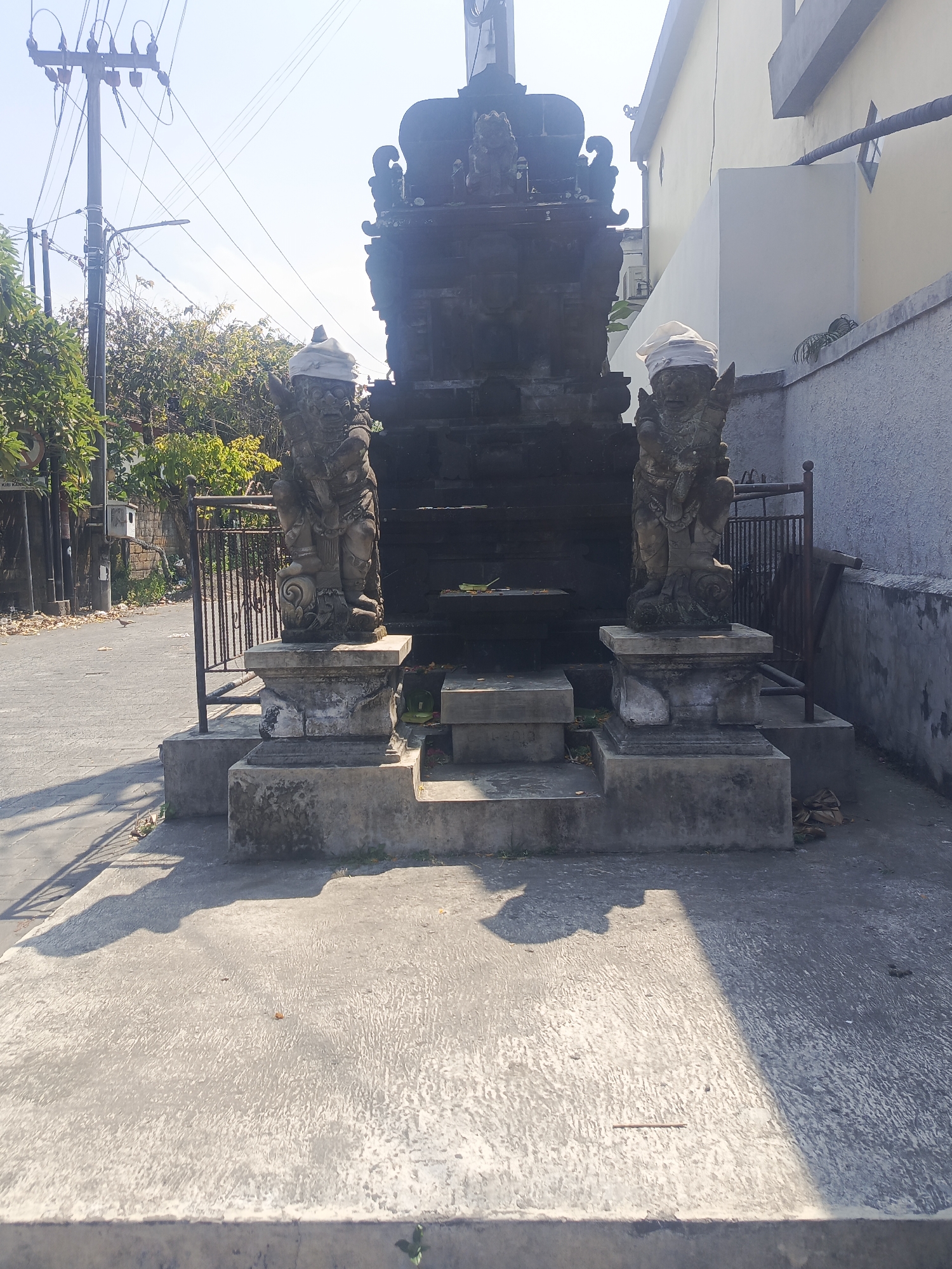 An outdoor, eye-level, medium shot captures a stone shrine with two guardian statues situated on a concrete paved area. The shrine is dark gray and appears to be made of stone, adorned with decorative carvings. The two stone statues, also dark gray, are positioned on either side of the shrine. Each statue is adorned with a white cloth wrapped around its head. Behind the shrine, a light yellow building with a gray lower section rises, and to the left, trees and a street with a utility pole are visible. The scene is bathed in sunlight, casting distinct shadows on the concrete. The ground is littered with a few fallen leaves.
