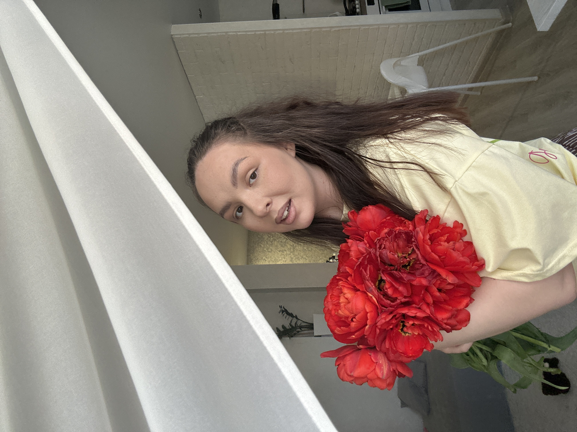 A woman with long brown hair, wearing a light yellow top, is holding a bouquet of red flowers with green stems. The flowers are in focus. She is looking at the camera. To the left of the woman is a white curtain. The background includes a white brick wall and a white chair. A dark object is visible on the floor near the base of the bouquet's stems. The location is likely in Chaikovsky, Russia.