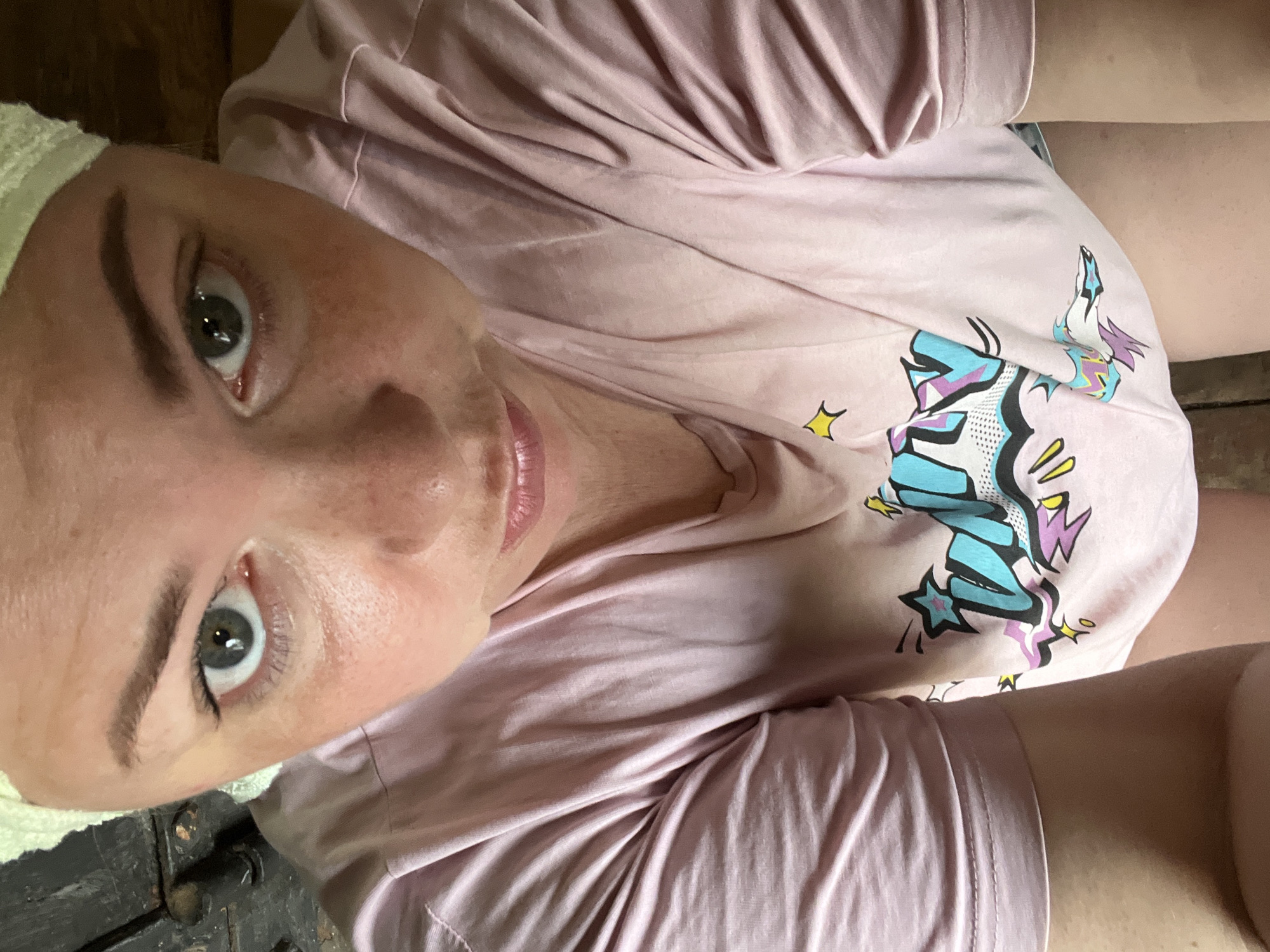 A woman with green eyes and fair skin, wearing a light pink t-shirt with a graphic print, is looking up at the camera. She has a light-colored headscarf on and is positioned in what appears to be an indoor setting with wooden elements in the background. The image is a close-up, focusing on her face and upper body, and does not provide any information about her location in the city of Fokino, Russia.