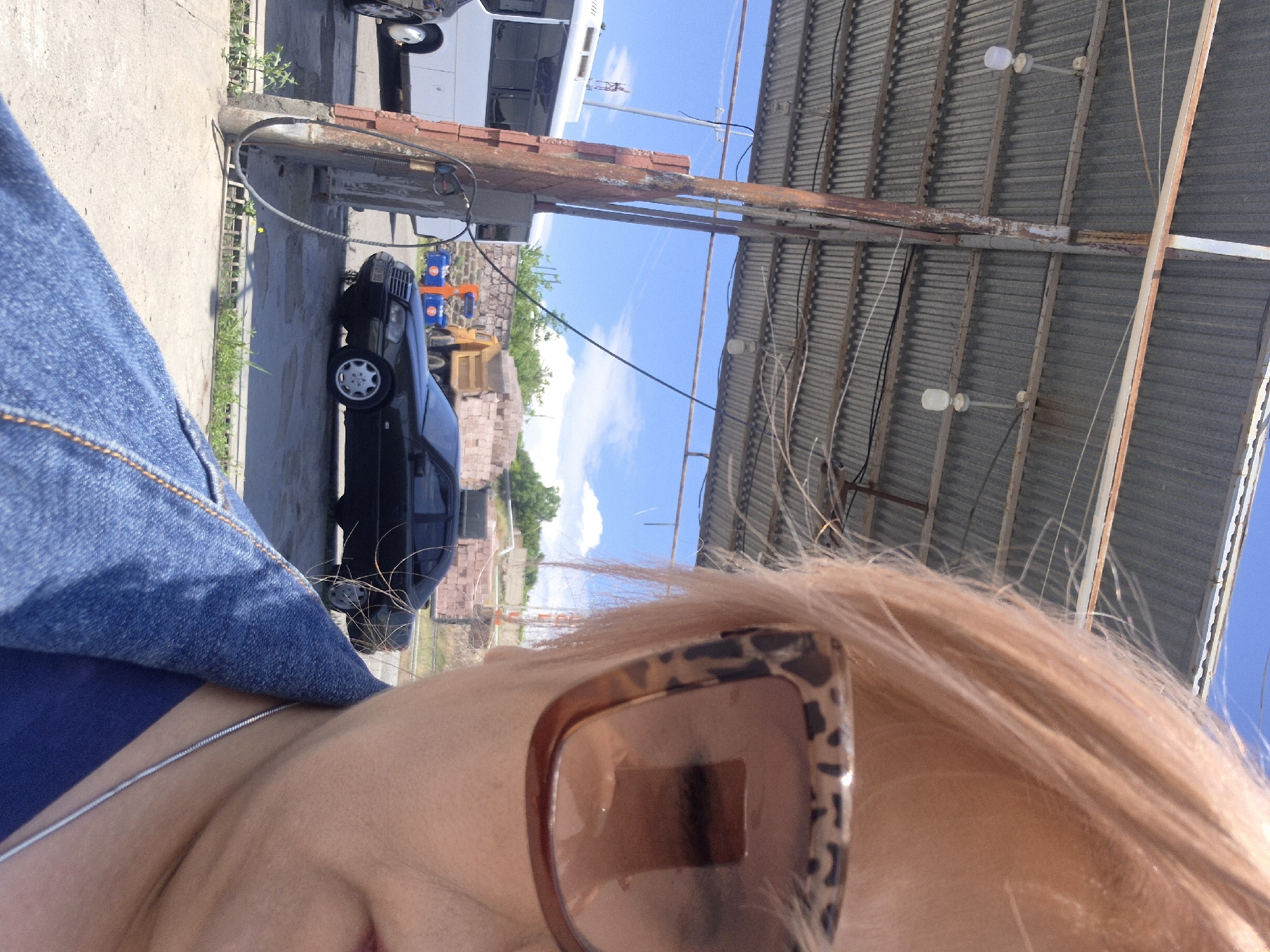The image is a close-up, selfie-style shot taken outdoors, likely at a gas station in Ashtarak, Armenia. The foreground is dominated by the upper portion of a person's face, wearing leopard-print sunglasses and a denim jacket over a blue shirt. The person's blonde hair is windswept.
In the background, a corrugated metal roof structure, supported by rusty metal poles and beams, is visible. Two light fixtures hang from the ceiling. Beneath the roof, a black Mercedes-Benz sedan is parked, and to its left, a white bus is parked. A yellow dump truck with a load of bricks is visible further in the background, behind the Mercedes. The environment appears to be a service area or industrial yard, with a paved ground surface. A power line is strung across the scene. The sky is blue with some scattered clouds. The presence of the bus, cars, and potential service infrastructure suggests a public or commercial area.