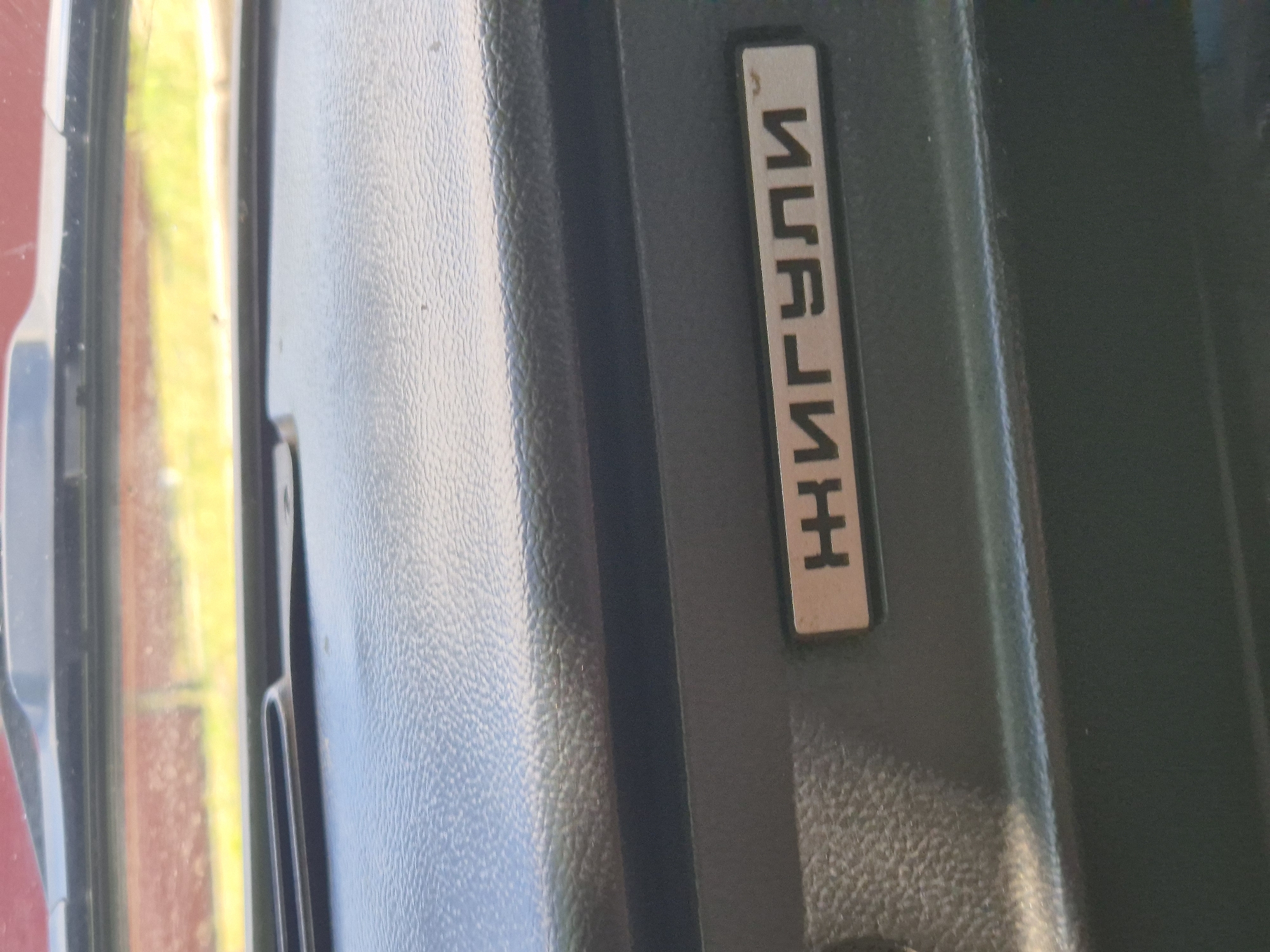 A close-up, eye-level shot shows the interior of a vehicle's dashboard. The foreground is dominated by a textured, dark gray surface, likely plastic. A metallic plaque is embedded in this surface, featuring Cyrillic lettering that reads "ЖИГУЛИ". The plaque is rectangular with rounded corners and appears to be made of brushed aluminum or a similar metal. Above the plaque, a slightly raised, textured area of the dashboard is visible. The background shows a blurry view through the car's windshield, revealing a predominantly yellow and brown landscape, suggesting an outdoor environment. The overall impression is of a well-worn vehicle interior. This image was taken in the village of Sozonivka, Ukraine.