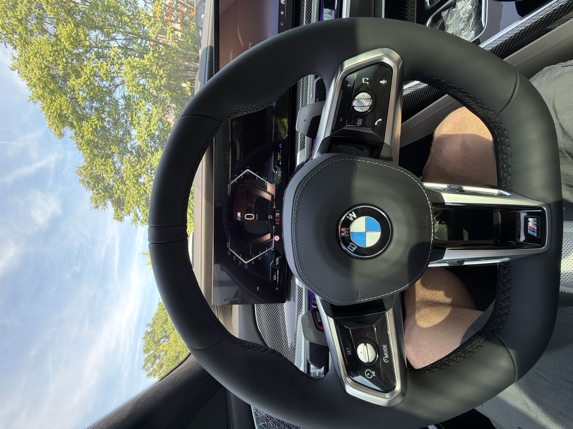 A first-person perspective shot from inside a BMW car shows the steering wheel and dashboard. The steering wheel is black leather with silver accents and the BMW logo in the center. The dashboard features a digital display showing speed, gear, and other vehicle information. A person's legs are visible below the steering wheel. Outside the car, green trees and a blue sky with clouds are visible. The location is Burgas, Bulgaria.