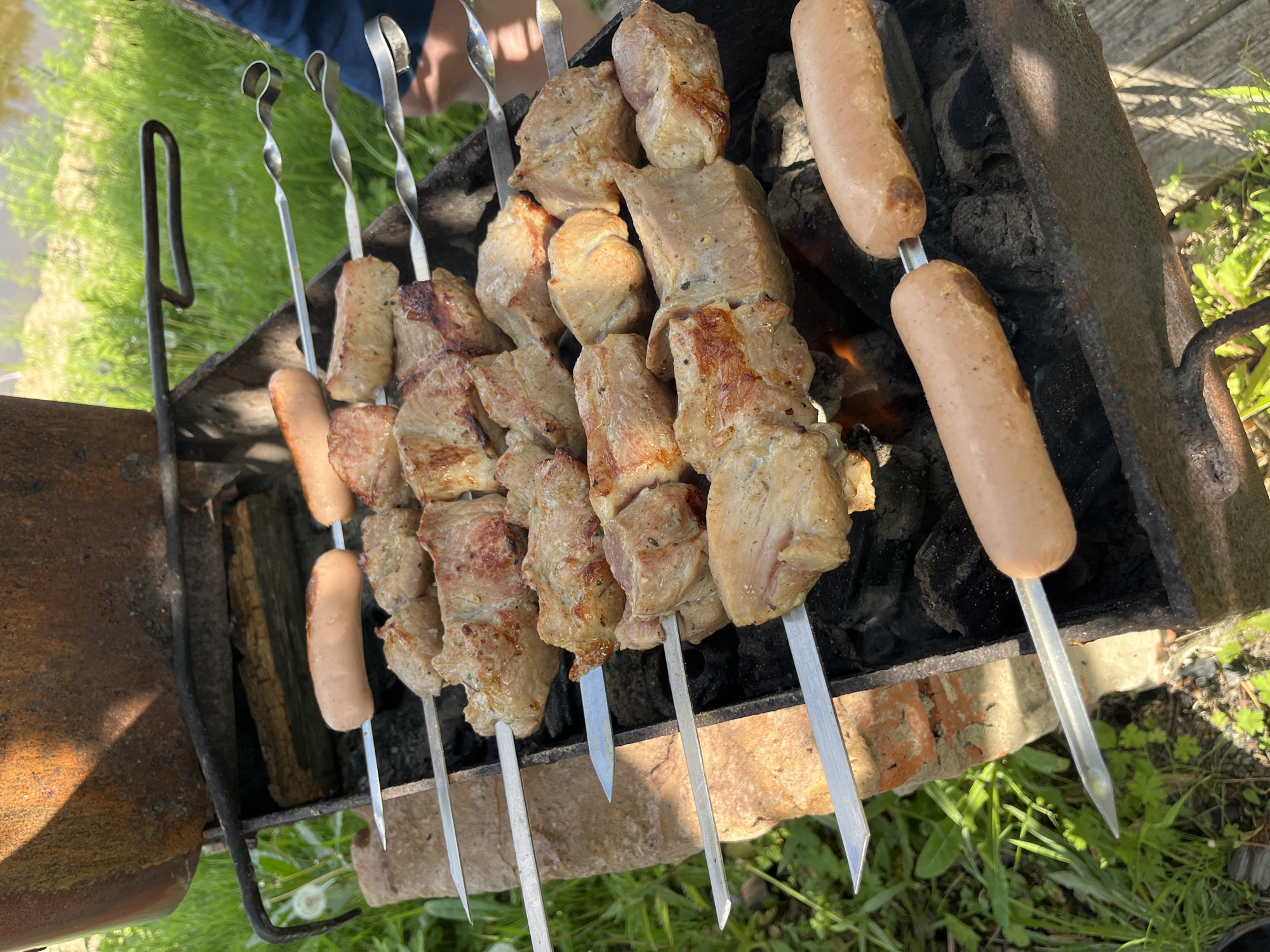 In Маяк, Russia, a close-up shot captures a barbecue with skewers of meat and sausages cooking over charcoal. The scene is set outdoors, with lush green grass and a body of water visible in the background, suggesting a natural or park-like setting. The barbecue grill itself appears to be made of metal and brick, with remnants of previous fires evident in the charred coals. The meat, cut into cubes, is skewered and shows signs of being grilled, with some pieces lightly browned and others more deeply seared. Several sausages, also on skewers, are positioned near the meat, indicating a mixed grill. The overall atmosphere is casual and suggests a leisurely outdoor meal or gathering.
