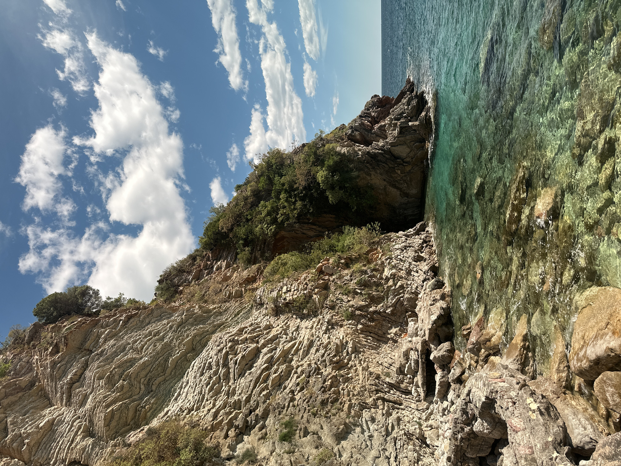 This image captures a serene coastal landscape under a bright blue sky dotted with scattered white clouds. A rugged cliff face, extending from the left and center, dominates the scene, meeting the clear, turquoise-to-emerald sea on the right. The lower sections of the cliff exhibit distinct, light tan to grey layered rock formations, displaying intricate wavy and folded patterns, suggesting geological activity. The upper parts of the cliff are densely covered with vibrant green vegetation, including shrubs and small trees. At the base of the cliffs, where they merge with the water, are rocky outcroppings and small natural alcoves. The exceptionally clear water reveals rocks and the seabed beneath its surface, transitioning from shallow clarity near the shore to a deeper blue towards the horizon. The scene depicts a peaceful daytime setting, with no human activity or visible text. The natural beauty of the rocky coastline, lush vegetation, and transparent sea is the focus. The location is Kamenovo, Montenegro, known for its picturesque Mediterranean coast.