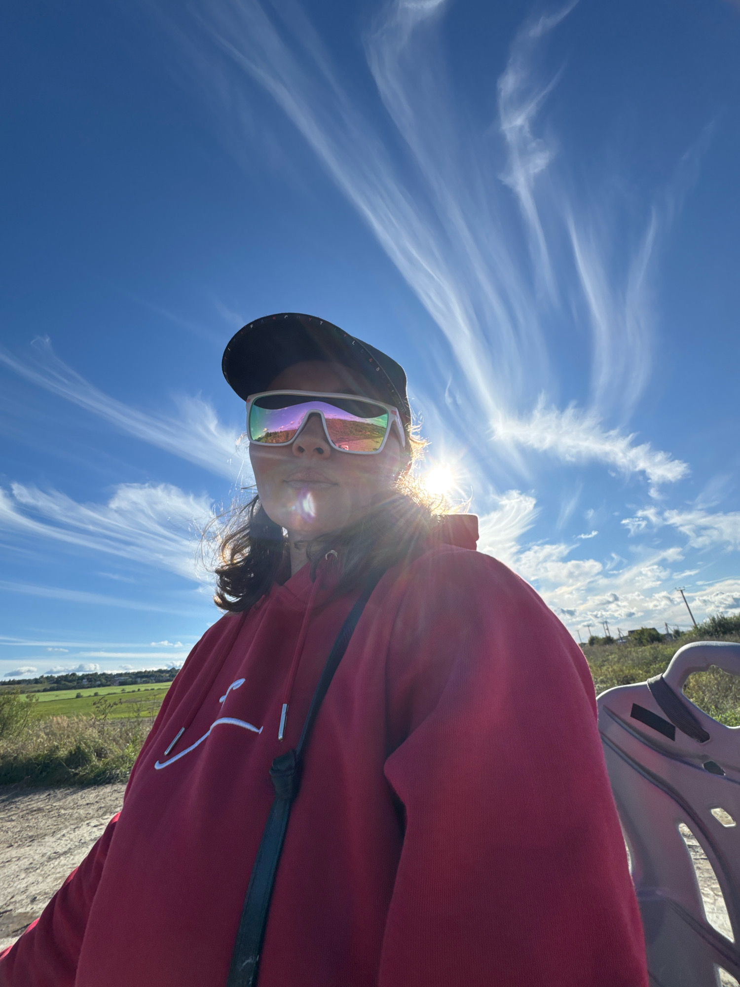 A low-angle, outdoor selfie captures a person wearing a red hoodie, a black baseball cap, and reflective sunglasses against a bright blue sky with wispy white clouds. The sun is visible behind the person, creating a lens flare effect. The person is seated, with the armrest of a light gray plastic chair visible on the right side of the frame. In the background, a rural landscape with green fields and distant trees is visible under the expansive sky. The scene appears to be in Kempelelevo, Russia.