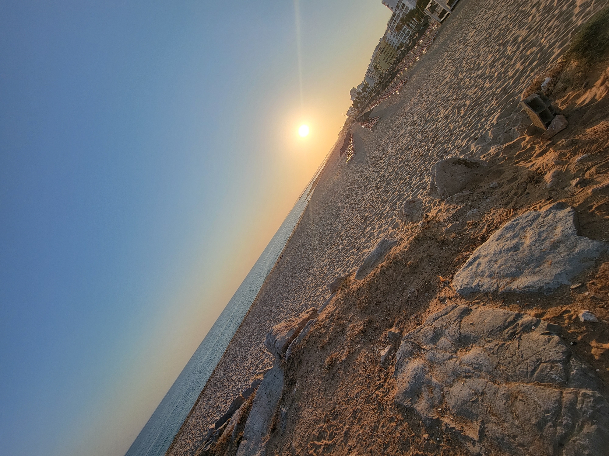 The image depicts a sun-drenched beach with the sun low on the horizon, casting a warm glow. The scene is angled, with the beach extending diagonally across the frame. The foreground features sandy terrain with scattered rocks and some dry vegetation. Further back, the expansive sandy beach is lined with rows of empty lounge chairs and umbrellas, indicating a resort area. Buildings, possibly hotels or apartments, are visible in the distance, suggesting an urban or semi-urban coastal location. The ocean is visible to the left, meeting the sky at a soft, hazy horizon. The sky itself transitions from a pale orange near the horizon to a clear blue overhead. The sun appears as a bright, luminous orb, with a visible lens flare to its right. The overall impression is one of a tranquil, end-of-day beach scene in Quarteira, Portugal.