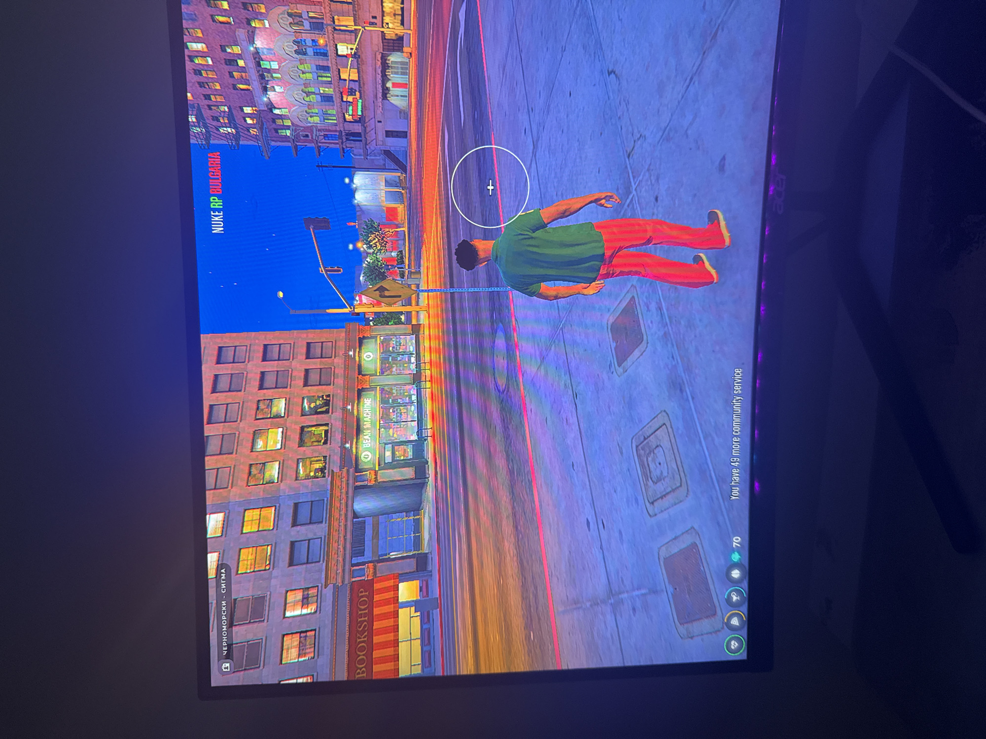 The image shows a video game scene. A male character wearing a green shirt and red striped pants stands in a city intersection. A white circle with a crosshair is in front of him, indicating a target or aiming reticle. In the background, there are buildings with illuminated windows, streetlights, and traffic lights. A sign at the top of the screen reads "NUKE RP BULGARIA". Another sign in the lower left corner shows "BOOKSHOP" and "BEAN MACHINE". A text box in the lower right corner states "You have 49 more community service". The scene depicts a virtual environment, likely from a role-playing game, set in a fictionalized Bulgarian city.