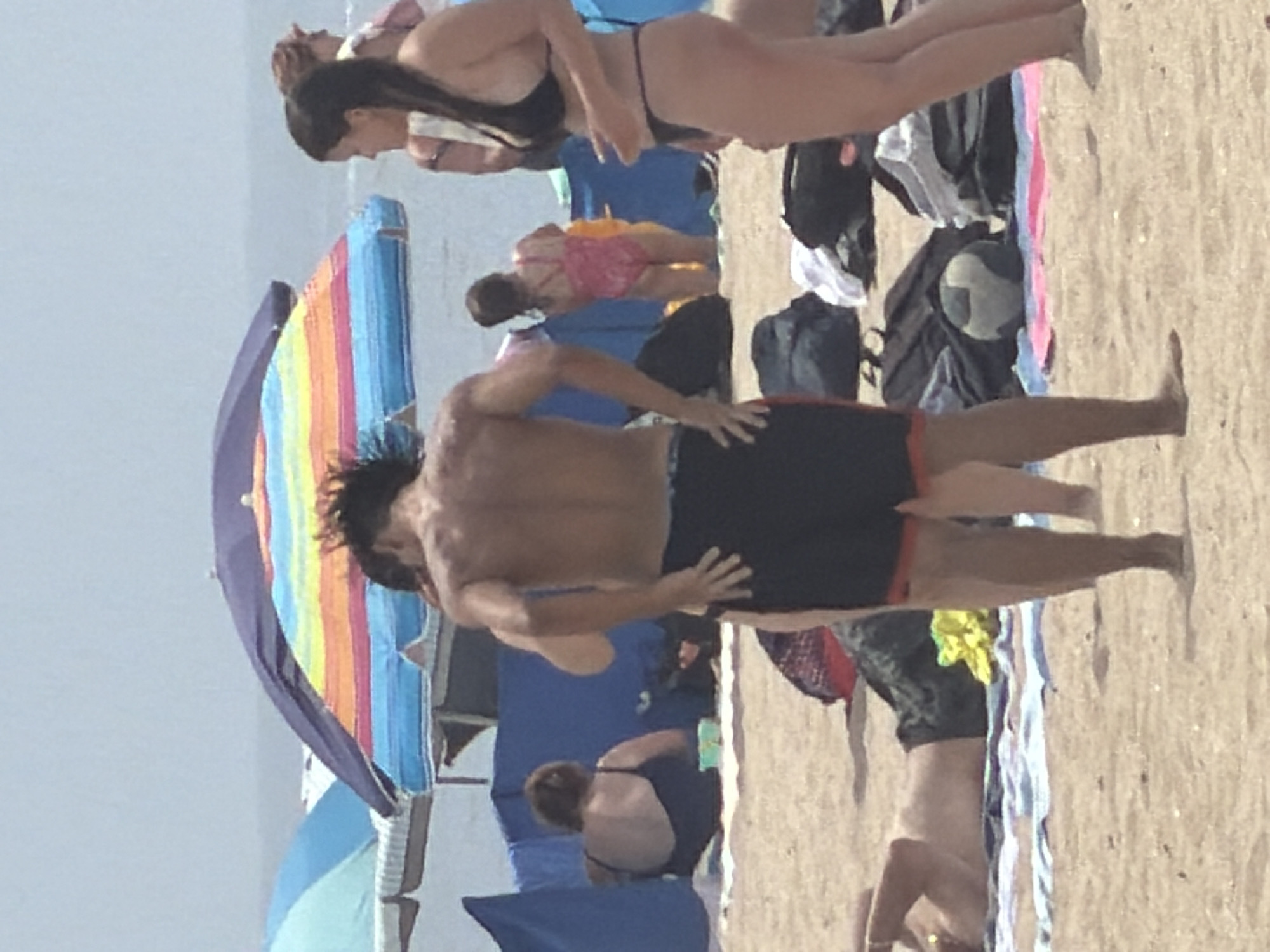 An outdoor, eye-level medium shot captures a beach scene in Espinho, Portugal, under a bright, hazy sky. In the foreground, a man with dark hair and a muscular physique, wearing dark swim trunks, stands with his back to the camera, facing the ocean. His left arm is bent at the elbow, and his left hand is resting on his hip. To his right, a woman with long dark hair, wearing a black bikini, is partially visible from the side. Her head is turned away from the camera, and her hands are clasped in front of her. In the background, a person in a pink swimsuit is visible. Further back, another individual, viewed from the back and wearing a dark swimsuit, sits on the sand. Two beach umbrellas are prominent; one is a large rainbow-colored umbrella, and another is a solid blue umbrella. Bags and towels are scattered on the sand, and a patterned beach ball rests on a blanket. The ground is composed of sand, and the ocean is visible in the distance. The overall atmosphere is that of a typical summer day at the beach.