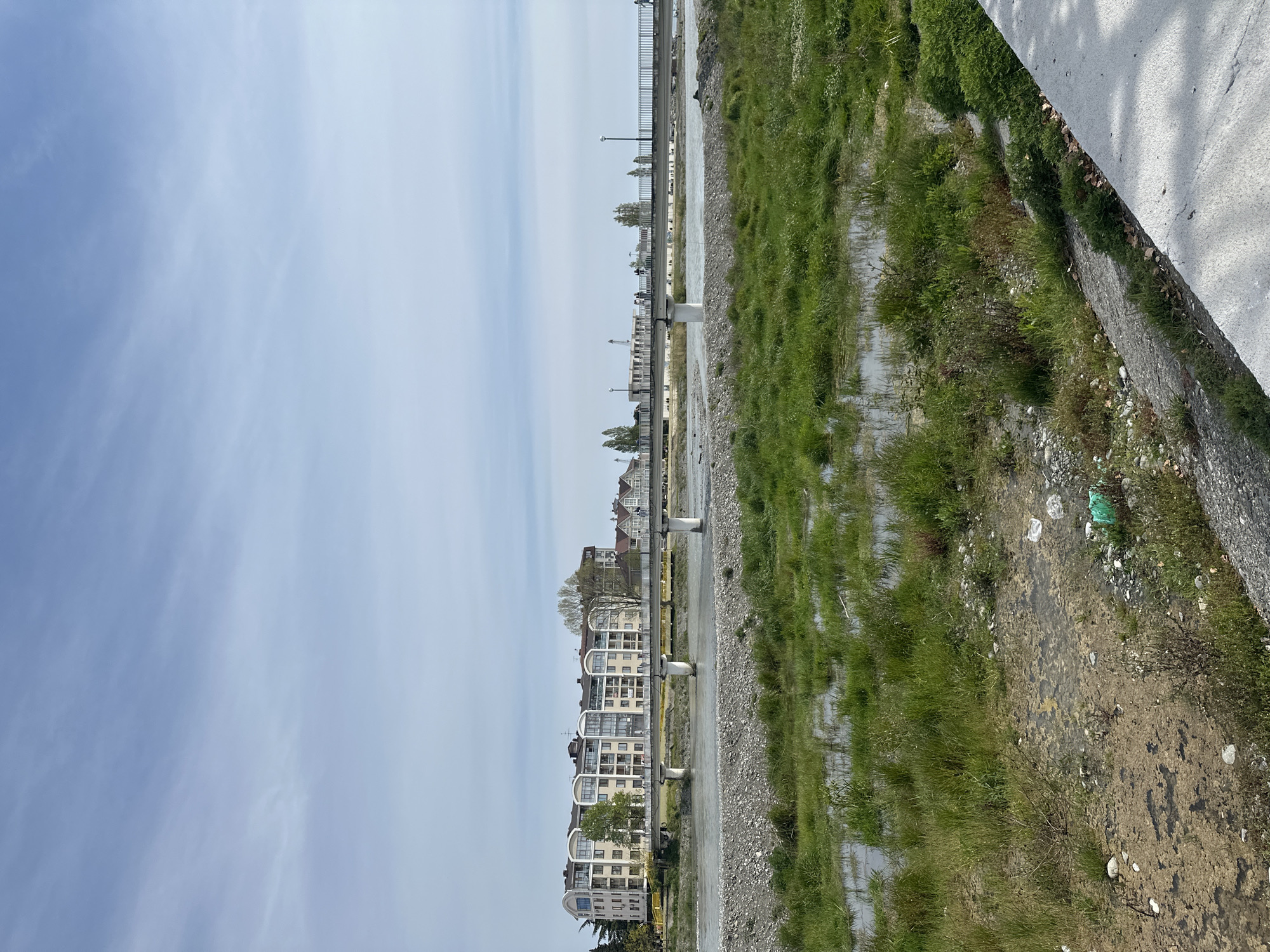 The image presents a wide, slightly elevated view overlooking a riverbed or channel in an urban or semi-urban setting, likely in Sochi, Russia. The perspective suggests it was taken from a bridge or an elevated walkway.
In the mid-ground, a broad, mostly dry riverbed dominates the scene. Patches of light blue water are visible, interspersed with areas of exposed gravel, dirt, and vibrant green grass growing in clusters and strips. This indicates low water levels or a seasonal flow.
On the left side of the riverbed, a modern, multi-story residential or hotel complex stretches horizontally. The buildings feature light-colored facades and distinct arched balconies, contributing to a contemporary architectural style. Parallel to these buildings and the channel, a pedestrian walkway or path is visible, supported by concrete pillars. Several streetlights are spaced along this path. Beyond these structures, more buildings are faintly discernible in the distant urban landscape.
The right side of the foreground features a large, light-colored concrete structure, possibly part of the bridge from which the photo was taken or an embankment wall. Shadows are cast upon this concrete surface, suggesting bright sunlight. Near the base of this concrete structure, along the riverbank, green vegetation grows, and some litter, including a noticeable green plastic bag, is scattered among the rocks and grass.
The sky above is mostly clear with a soft blue hue, showing only very light, wispy clouds. The lighting suggests it is daytime, likely morning or afternoon. No people or specific activities are visible, presenting a calm, static landscape. There is no visible text in the image.