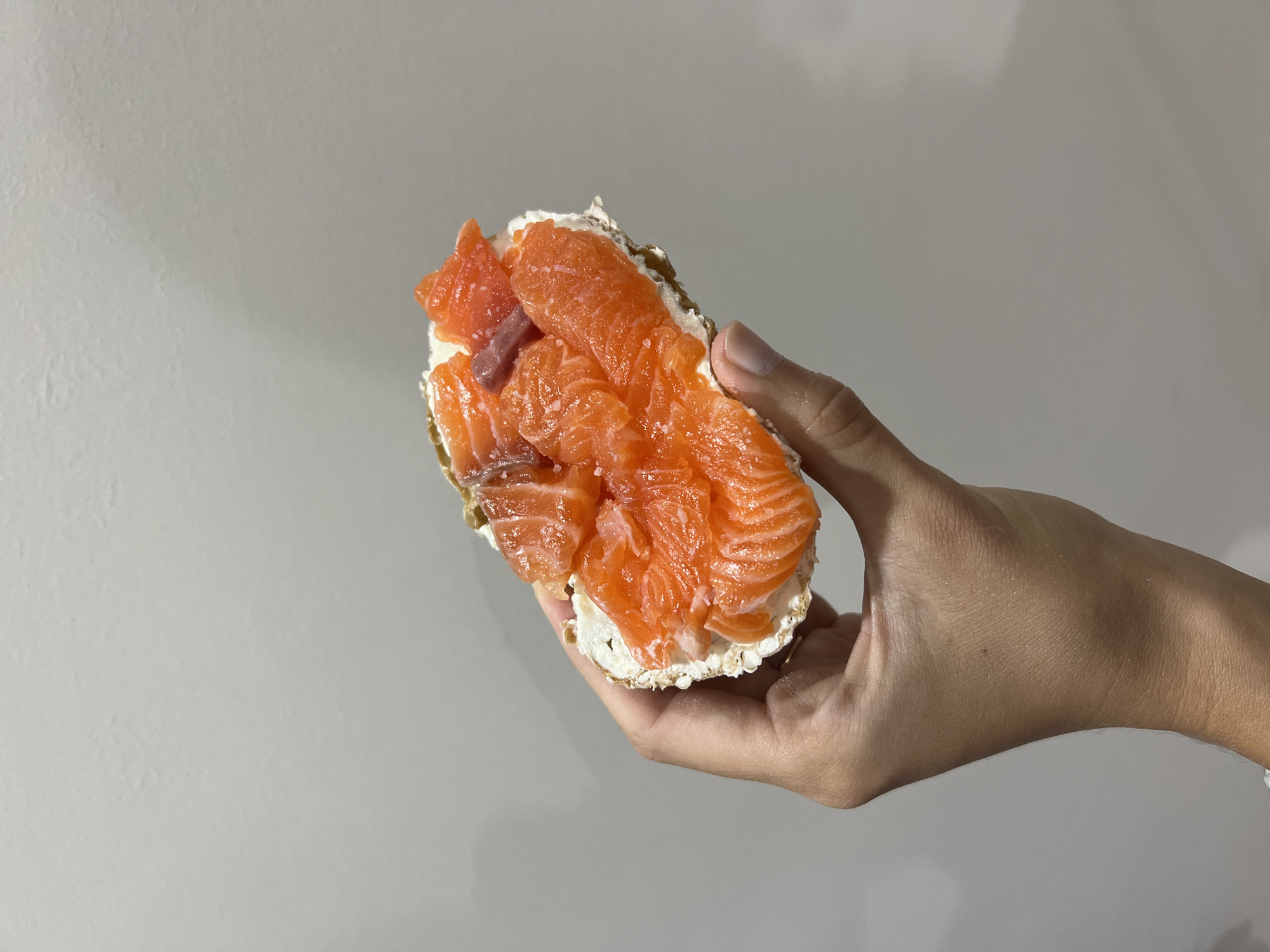 A hand holds a bagel with cream cheese and salmon. The background is a plain, light gray wall. The scene appears to be indoors. It is not possible to confirm the location based on the image.