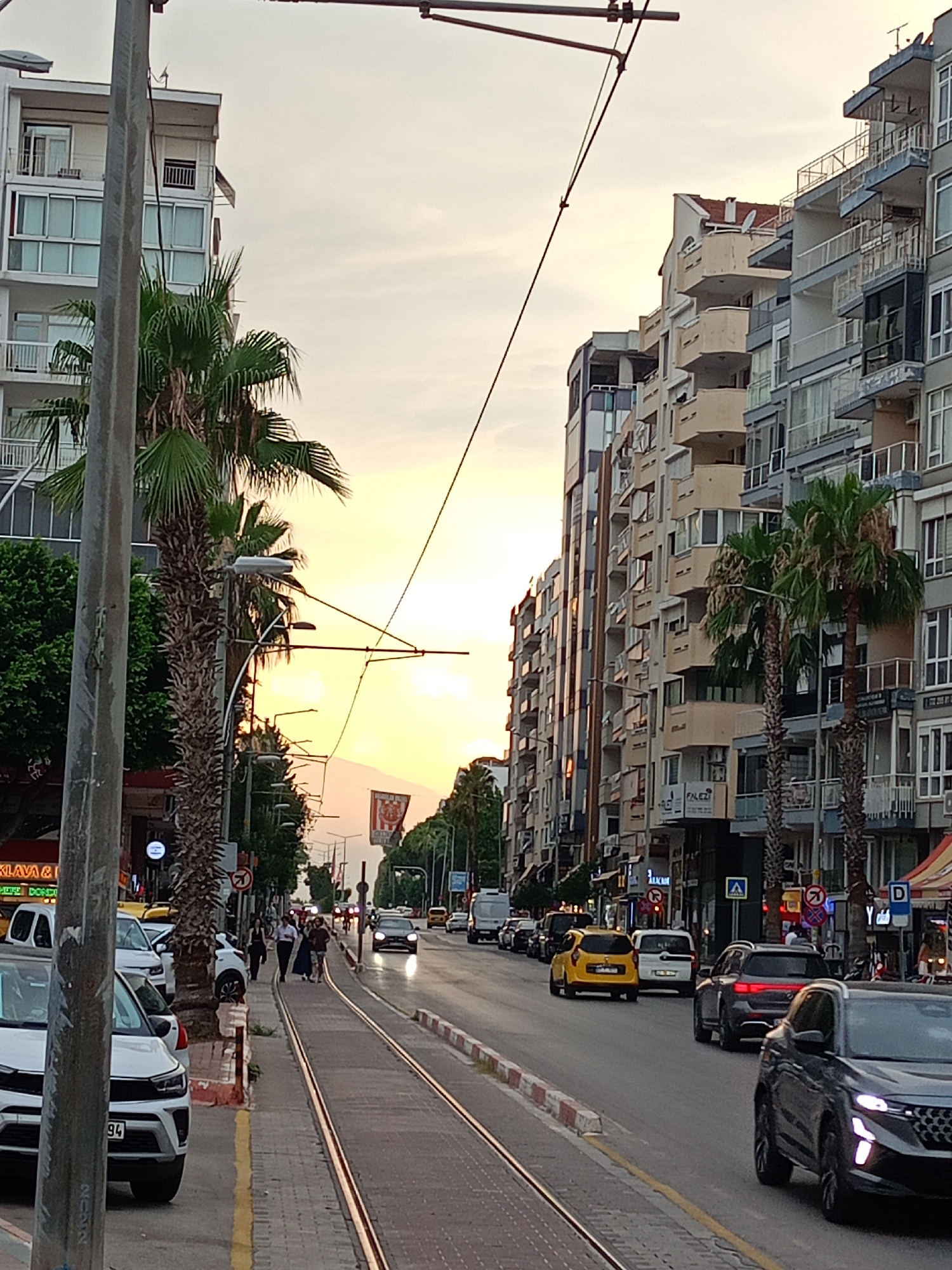 The image depicts a street scene in Antalya, Turkey, at sunset. The foreground features a concrete utility pole and tram tracks running along the side of the road. On the left, a white car is partially visible, and behind it, a multi-story building with balconies and windows stands, with a lush palm tree in front.
The street itself is lined with buildings on the right, characterized by their modern architecture, balconies, and numerous windows. These buildings appear to be residential or commercial complexes. Palm trees are scattered along the sidewalk, adding to the Mediterranean ambiance.
Several cars are present on the road, including a yellow taxi and other vehicles in motion. Pedestrians are walking on the sidewalk, and some are crossing the street. The sky above is filled with the warm hues of sunset, with a bright yellow and orange glow, indicating the sun is low on the horizon. A banner with a red and white pattern is visible in the distance, hanging from a pole. The overall scene suggests a bustling urban environment during a pleasant evening.