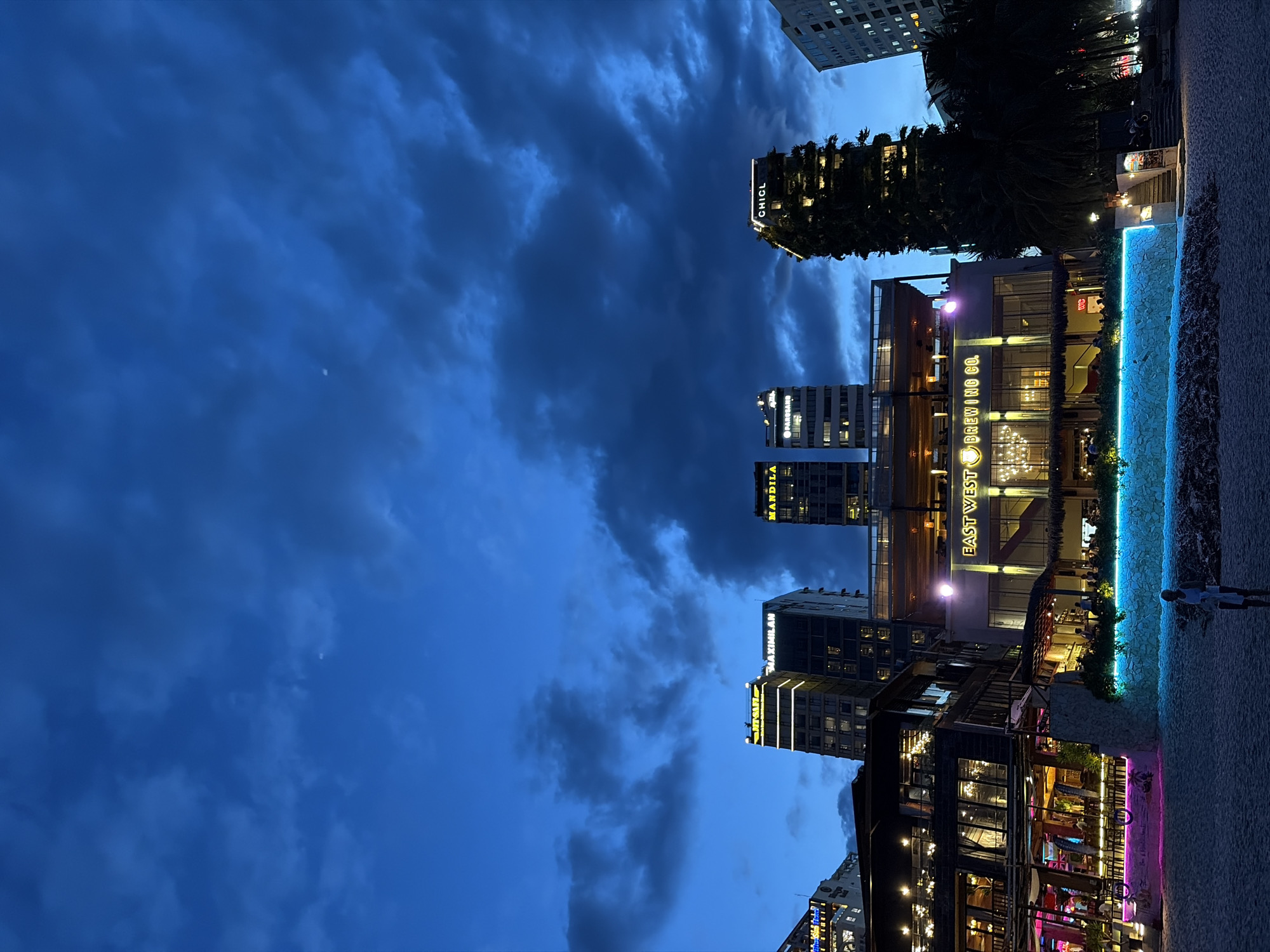 This image depicts a twilight scene on a beach in Đà Nẵng, Vietnam. The sky is a deep blue, filled with dark, dramatic clouds. In the background, several modern buildings rise, some illuminated with neon lights and signage. Notable buildings include "MAXIMILAN" and "MANDILA," with another building identified as "CHICL" that is covered in greenery and illuminated from within.
In the midground, a multi-story establishment with a broad facade is visible, featuring large windows and a prominent sign reading "EAST WEST BREWING CO." The building is warmly lit from within, and outdoor seating areas are apparent on multiple levels. A decorative wall in front of this establishment glows with vibrant blue and pink neon lights. The foreground shows a sandy beach. A single person, a man wearing a white shirt and shorts, stands on the sand, facing away from the camera and towards the ocean. Beach chairs and other amenities are hinted at on the left side of the frame. The overall atmosphere is that of a lively coastal urban setting transitioning from day to night.