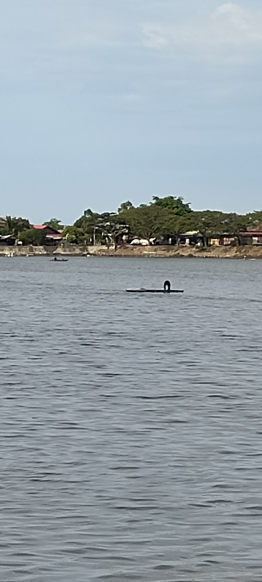 The image shows a wide body of water, likely a river or bay, with a shoreline in the background. On the water, there is a dark object with a curved section, possibly a submerged structure or debris. In the distance, on the shore, there are buildings and lush green trees. There are also two small boats with people in them on the water. The sky is a pale blue with some scattered clouds. The overall scene depicts a serene waterfront environment in Lhokseumawe, Indonesia.