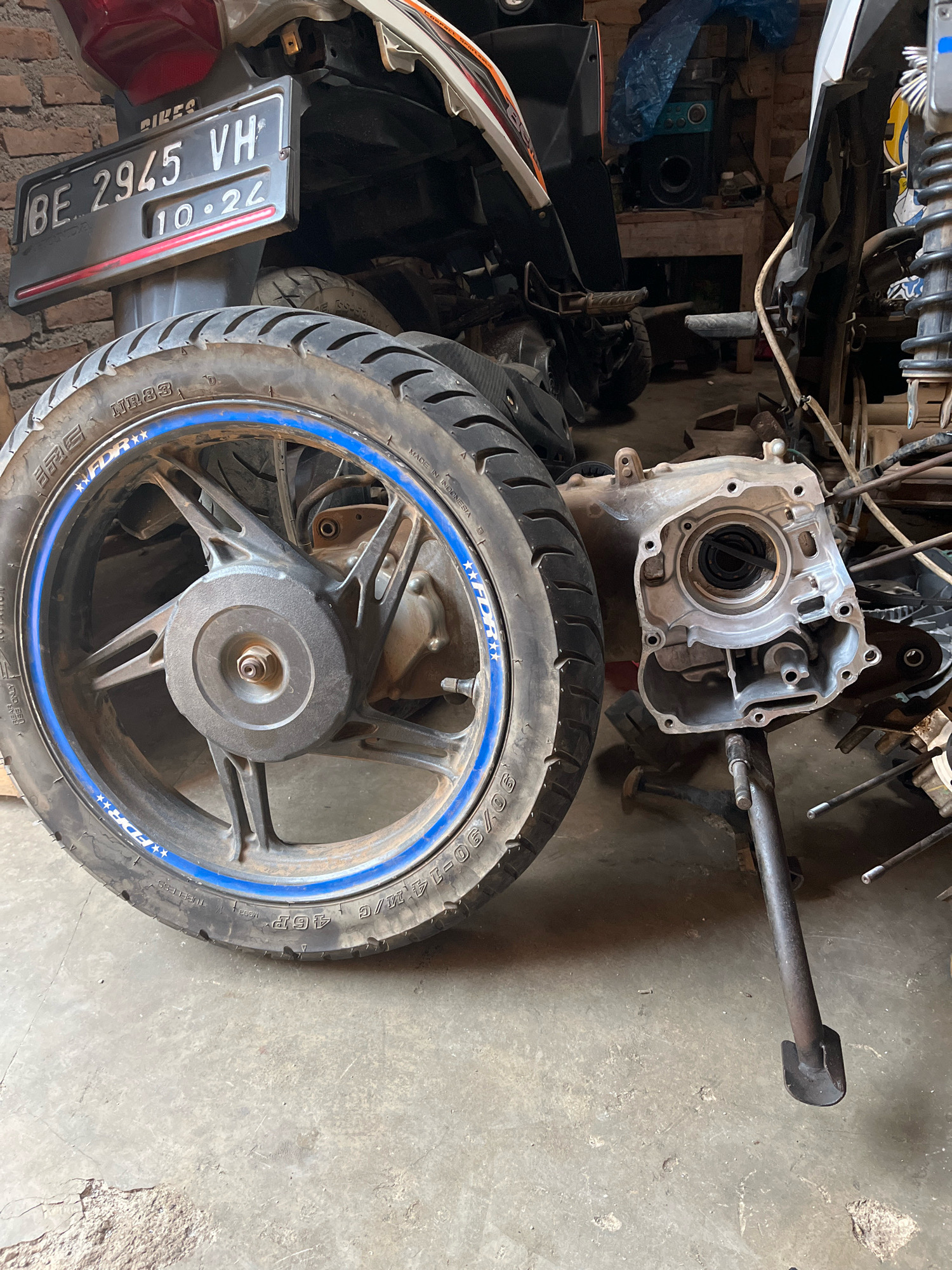 The image shows a disassembled motorcycle, likely a scooter, in a workshop or garage. The focus is on the rear wheel and a portion of the engine casing, which has been removed. The motorcycle has a license plate that reads "BE 2945 VH 10-24". The environment appears to be a concrete floor with brick walls in the background, suggesting an indoor setting. There are also other motorcycle parts and tools scattered around, indicating ongoing repair or maintenance work. The overall scene suggests a typical motorcycle repair shop or a personal workshop for vehicle maintenance. There are no people visible in the image. The scene is static and appears to have been taken during daylight hours.
