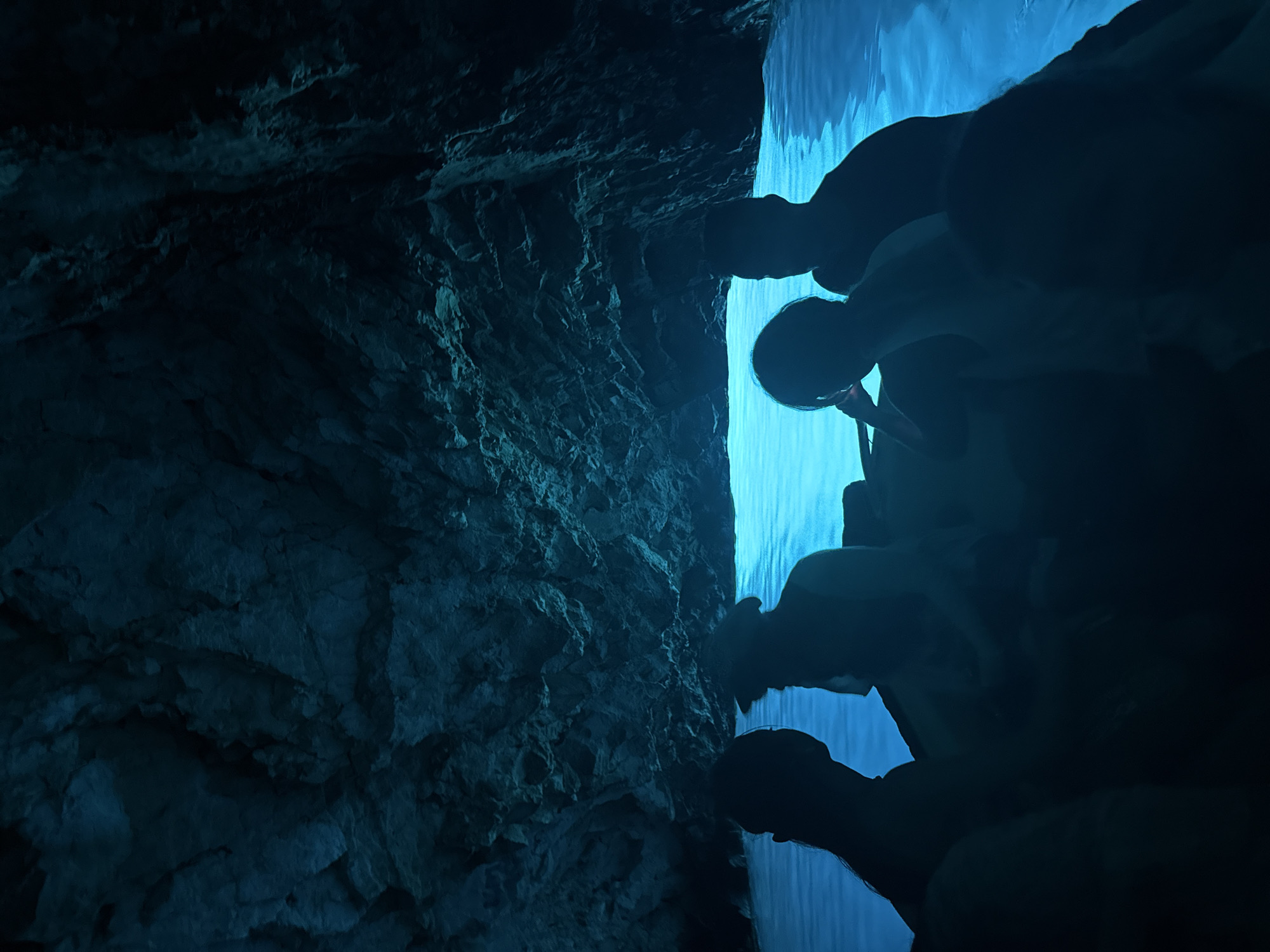 This image shows a group of people in a boat inside the Blue Cave, a sea cave on the island of Vis, near the town of Komiža, Croatia. The cave's interior is illuminated by a surreal, bright blue light, created by sunlight reflecting off the sandy seabed and passing through an underwater opening. The people are silhouetted against the glowing water, appearing as dark shapes within the dimly lit cave. The rocky walls of the cave rise steeply above the water, their rough textures visible in the faint light. The overall scene conveys a sense of mystery and wonder, with the vibrant blue water contrasting dramatically with the dark cave and its occupants.