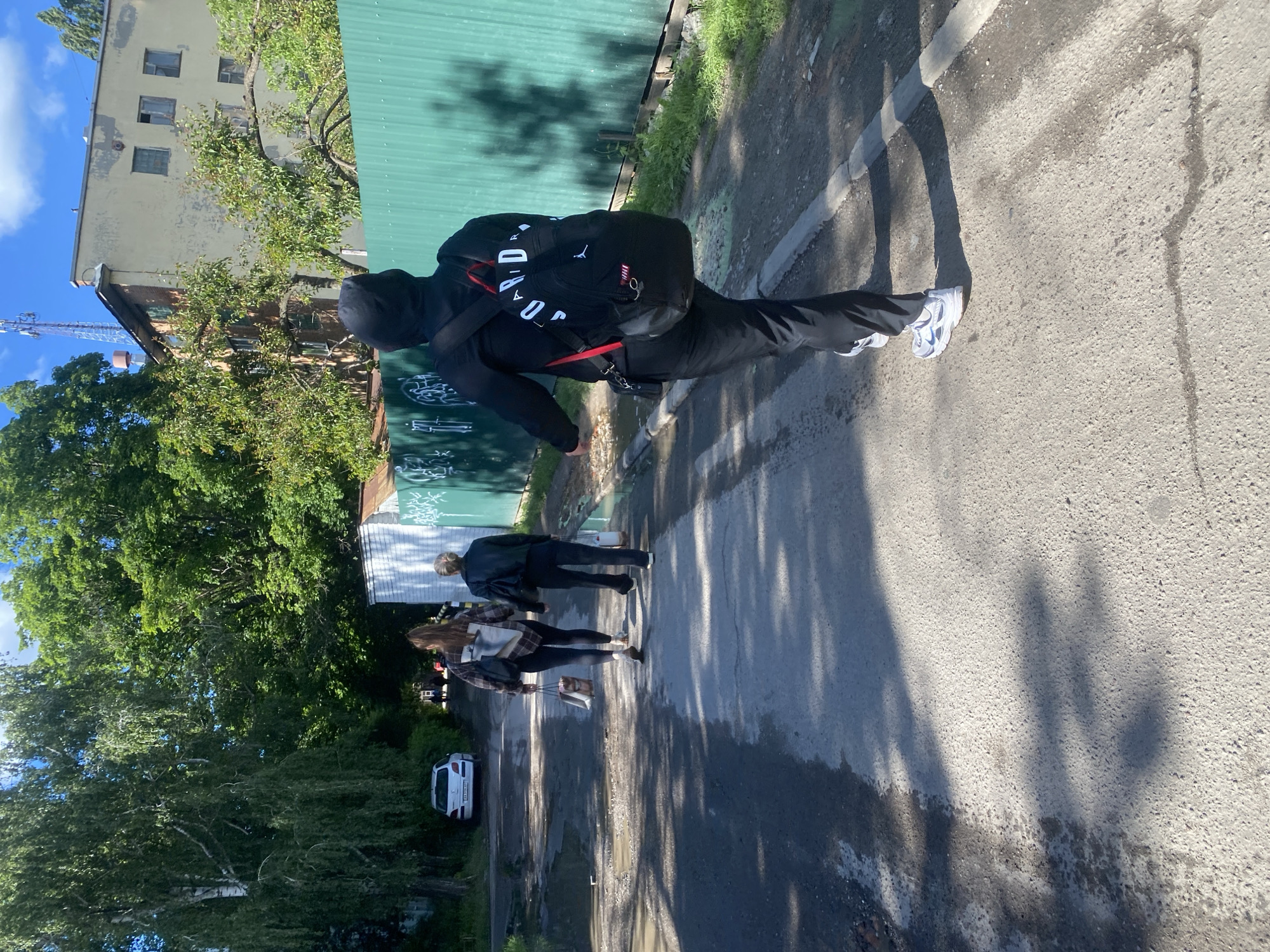 The image shows an urban scene in Poltava, Ukraine. In the foreground, a person wearing a black hooded sweatshirt, black pants, and white sneakers walks along a paved road, carrying a black backpack with red accents. Further down the road, two more people walk in the same direction. A white car is parked on the left side of the road. Green trees and vegetation line the left side of the road, while a tall building and a green corrugated metal fence are visible on the right. The sky is blue with some clouds. The road surface is slightly wet in places.