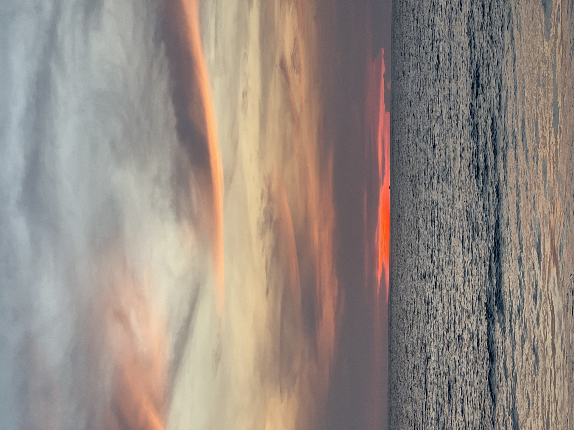 The image captures a vibrant sunset over water. The sky is filled with clouds, colored in shades of gray, yellow, orange, and red, indicating the setting sun. The water surface is textured with waves, reflecting some of the colors from the sky. It is currently unknown where in Unknown, Unknown, the photograph was taken.