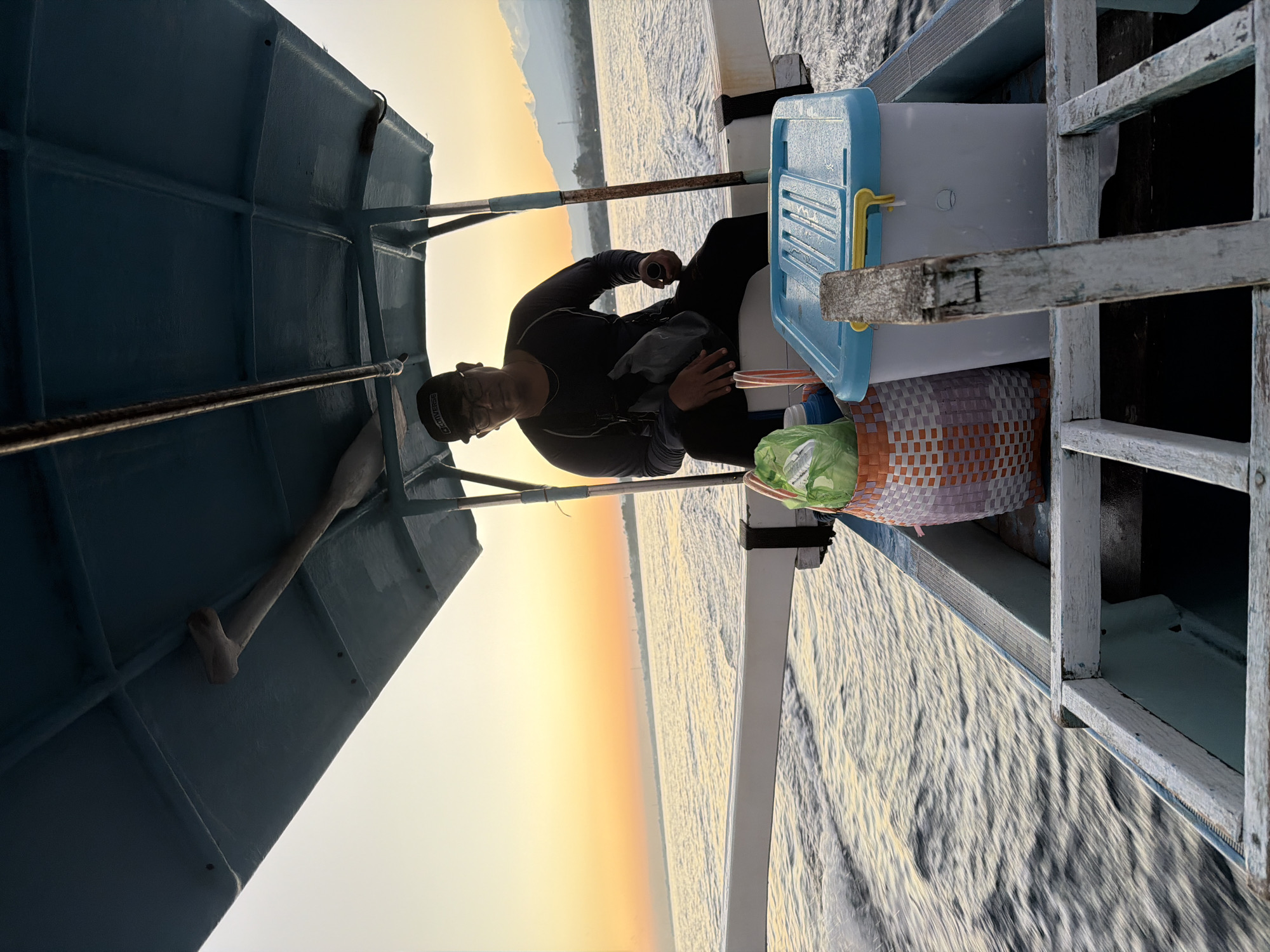 The image shows a man sitting in a small boat on the water, with a sunset in the background. The man is wearing a black long-sleeved shirt, black pants, and a black baseball cap. He is looking at the camera and holding a black bag. In the boat with him are a woven basket with green items inside, and a blue and white cooler. The boat appears to be moving through the water, creating ripples and waves. The sky is a warm orange and yellow, indicating it is either sunrise or sunset. In the distance, there is a hazy shoreline with mountains. The overall scene suggests a peaceful journey at sea, possibly near the coast of Banjar, Indonesia, given the tranquil water and the presence of mountains in the distance, which is characteristic of the region.