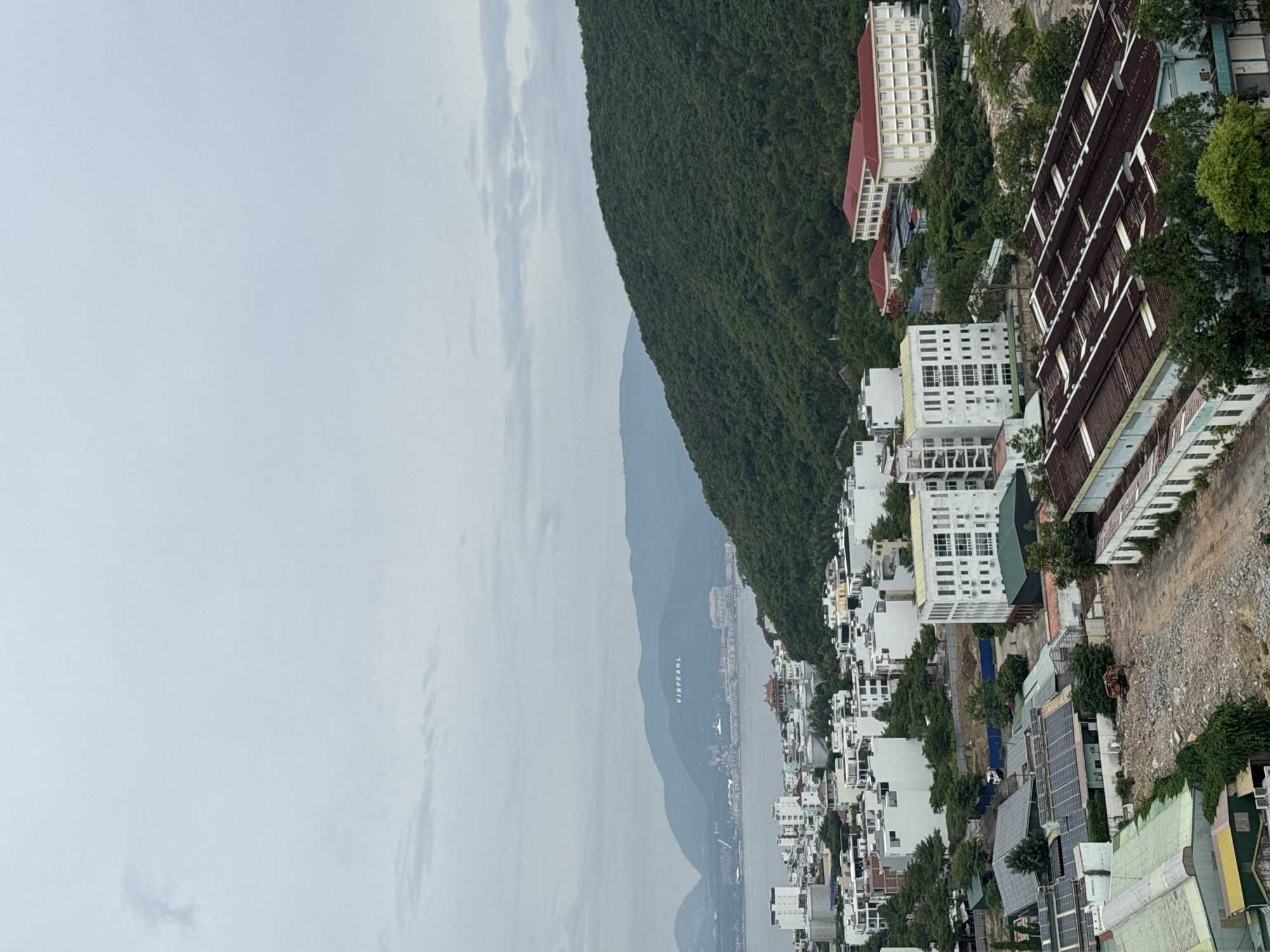 An aerial view of a coastal city, likely Nha Trang, Vietnam, under a cloudy sky. In the background, lush green mountains rise, with the word "VINPEARL" visible in large white letters on the slope of one mountain. A body of water, presumably the sea, separates the mountains from the city. The city itself is a dense collection of buildings, predominantly white and light-colored, of varying heights. Some buildings appear to be residential or commercial, with modern architecture featuring multiple windows and balconies. A few larger structures, like hotels or institutional buildings, with red roofs, are also present. In the foreground, there are industrial-looking buildings with corrugated metal roofs, some appearing aged and in disrepair. Construction or demolition is evident in some areas, with piles of rubble and construction equipment, including an excavator, visible. Trees and vegetation are interspersed throughout the urban landscape, particularly on the hillsides. The overall impression is a developing city situated in a picturesque natural setting.