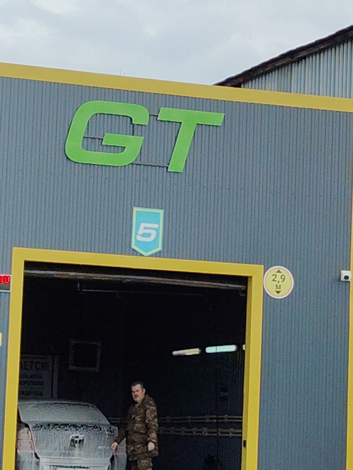 A medium shot depicts the exterior of a car wash, possibly in Unknown, Unknown. The structure is characterized by a corrugated metal facade in a muted blue, trimmed with bright yellow accents outlining the building and entrance. The signage above the entrance displays "GT" in a vivid green font, and beneath, a badge-like emblem with the number "5." To the right of the entrance, a circular sign indicates a height restriction of "2.9 M."
The open entrance reveals the dark interior of the car wash, where a car is undergoing cleaning, its surface covered in soap suds. A man in camouflage attire stands beside the vehicle.