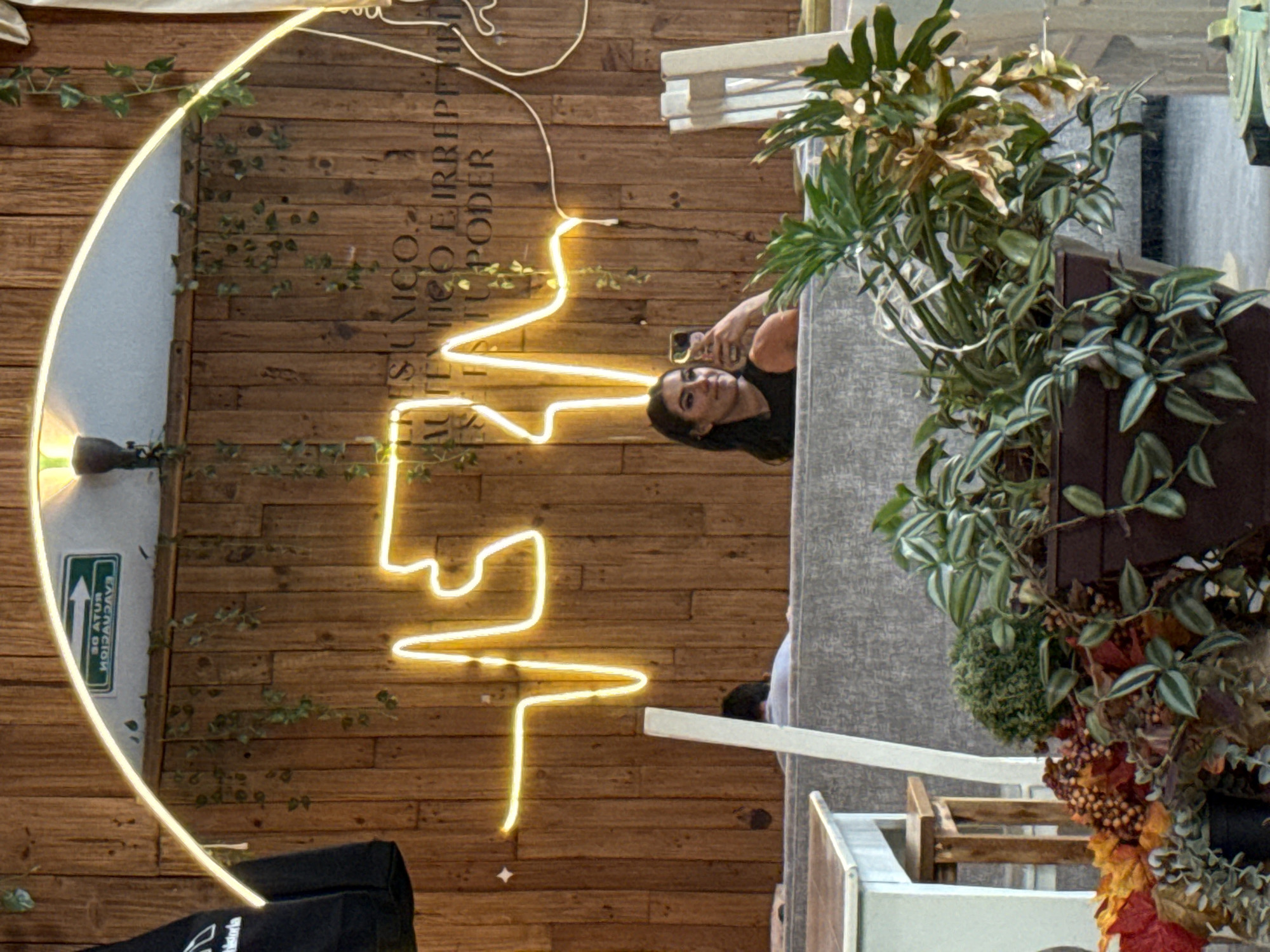The image shows a woman taking a picture of a mirrored art piece on a wooden wall. The art piece is a round mirror with a neon light sculpture, and the wall has green vines. The mirror reflects part of the text "ES SU UNICO E IRREPETIBLE PODER". The woman is holding a phone and facing the mirror. She has dark hair and a black shirt. To the right of the woman is a table with plants in pots. In the lower left corner is a black bag. The location is likely a restaurant in Puebla, Mexico, due to the unique architectural design and the sign in Spanish on the wall. The overall environment appears to be an indoor dining space with decorative elements.