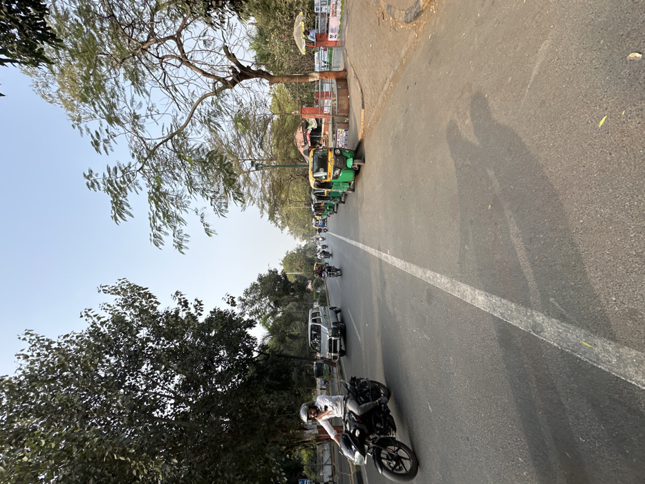 The image captures a daytime street scene in Vadodara, India, depicting a multi-lane asphalt road under a clear light blue sky. A faded white lane divider separates the road. On the left, a person wearing a white shirt and a light-colored helmet rides a black motorcycle, moving from left to right across the foreground. Behind the motorcycle, a silver van proceeds in the same direction, followed by additional distant motorcycles. On the right side of the road, a line of at least five green and yellow auto-rickshaws are parked parallel to the curb. Adjacent to these vehicles, a paved area features several small, reddish-brown structures resembling vendor stalls, one with a yellow awning, and another with a white umbrella covering a partially visible individual in a blue shirt. A low red brick wall or fence runs along this right-hand side, displaying partially visible signage. Dense green trees line both sides of the street, casting long shadows diagonally across the road from a light source to the left.