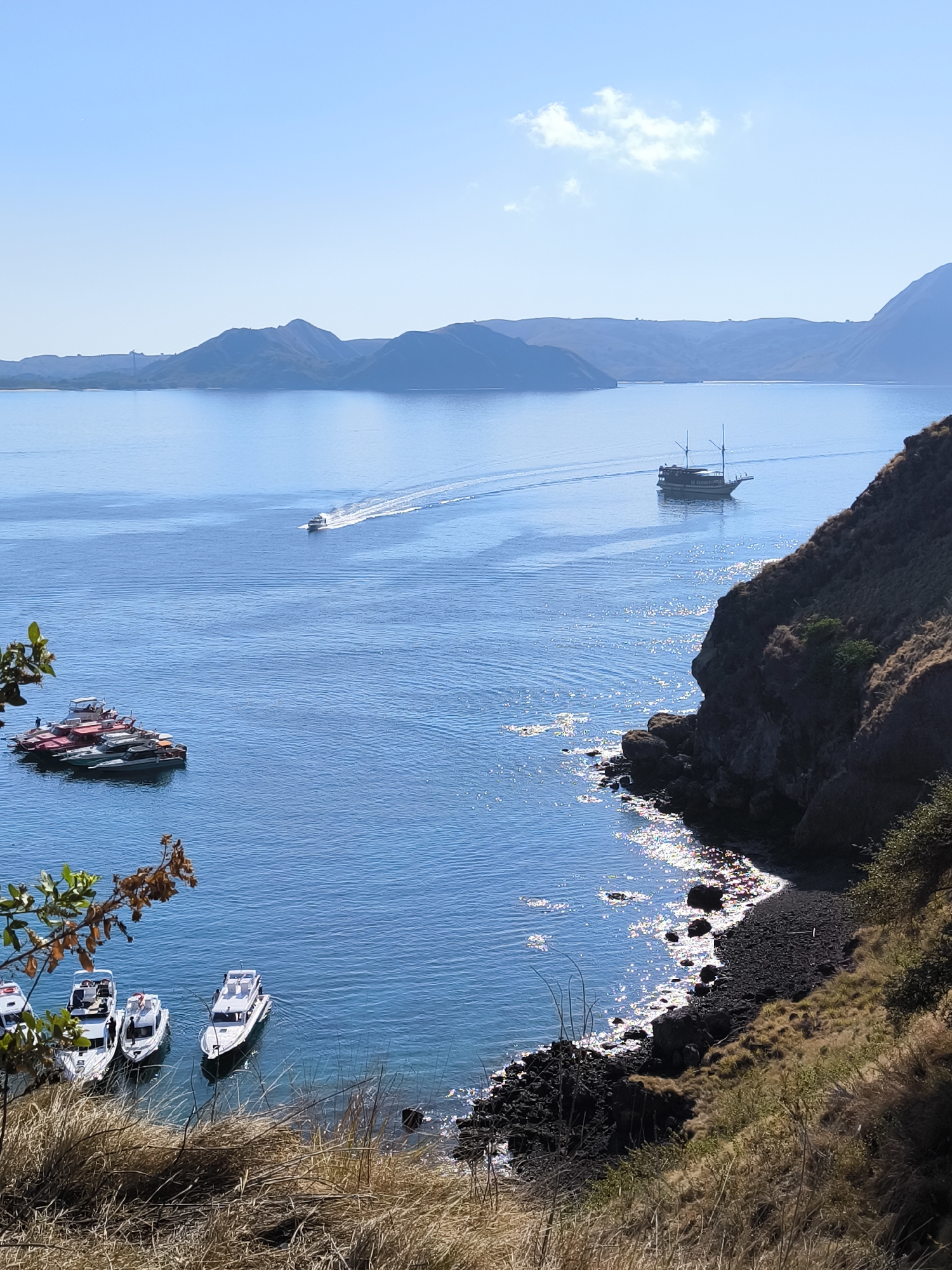 The image depicts a scenic coastal view with a clear blue sky and calm waters. In the distance, a chain of lush green hills borders the horizon. The sea is dotted with several boats of various sizes. To the left, a cluster of smaller boats is anchored together. Further out in the water, a medium-sized boat with two masts is sailing. In the mid-ground, a faster boat is creating a white wake as it speeds across the water. The right side of the image is dominated by a steep, rocky cliff face covered in dry, brown vegetation, sloping down to a black volcanic pebble beach. The sun glints off the water's surface, creating a shimmering effect along the coastline. In the foreground, dry grass and sparse foliage are visible.