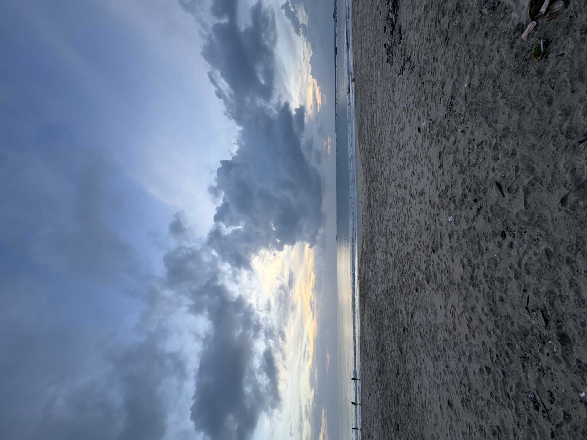 This image captures a serene yet dramatic beach scene at Kuta, Indonesia, likely around sunrise or sunset. The sky, occupying the upper two-thirds of the frame, is dominated by heavy, dark grey clouds, particularly on the left side, hinting at a recent storm or overcast conditions. Towards the right, the clouds break, revealing lighter patches and subtle glows of pale yellow and orange, indicating the sun's position near the horizon.
Below the sky, a calm ocean stretches across the midground, reflecting the subdued light. Gentle waves are visible near the shore, and several small, silhouetted figures can be seen in the distance, near the water's edge, possibly wading or engaging in activities like surfing.
The foreground consists of a wide expanse of dark, wet sand, textured with numerous footprints and small undulations. Scattered pieces of debris, including what appears to be wood and plastic, are visible on the sand, particularly in the lower right corner. The overall ambiance is one of quiet contemplation, with the shifting light and cloud formations creating a captivating natural spectacle. There is no visible text in the image.