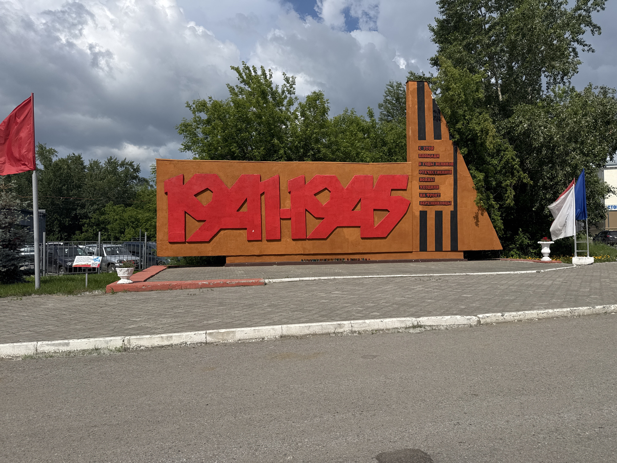 This is a medium-shot, eye-level, outdoor photograph of a monument in Usolye, Russia, dedicated to the years 1941-1945, symbolizing the Great Patriotic War. The monument is a large, orange, abstract structure with bold red numbers "1941-1945" prominently displayed on its front. To the right of the numbers, on a narrower, darker section of the monument, is a vertical inscription in Russian, with text including "В ЭТОМ МЕСТЕ" (In this place), "В ГОДЫ ВЕЛИКОЙ ОТЕЧЕСТВЕННОЙ ВОЙНЫ" (In the years of the Great Patriotic War), and "УХОДИЛИ НА ФРОНТ БЕРЕЗНИКОВЦЫ" (Bereznikovtsy went to the front).
In the background, to the left of the monument, a tall flagpole flies a red flag. Behind the monument, a line of lush green trees and foliage forms a natural backdrop. To the right of the monument, two flags are visible: a white flag and a blue and white flag, possibly the Russian or a regional flag. The foreground consists of a paved walkway made of grey interlocking bricks, bordered by a red curb. Beyond the walkway, a gray asphalt road is visible. Several parked cars and a fence are discernible in the far background on the left. The sky is partly cloudy with patches of blue sky and gray clouds. The overall lighting suggests it is daytime.