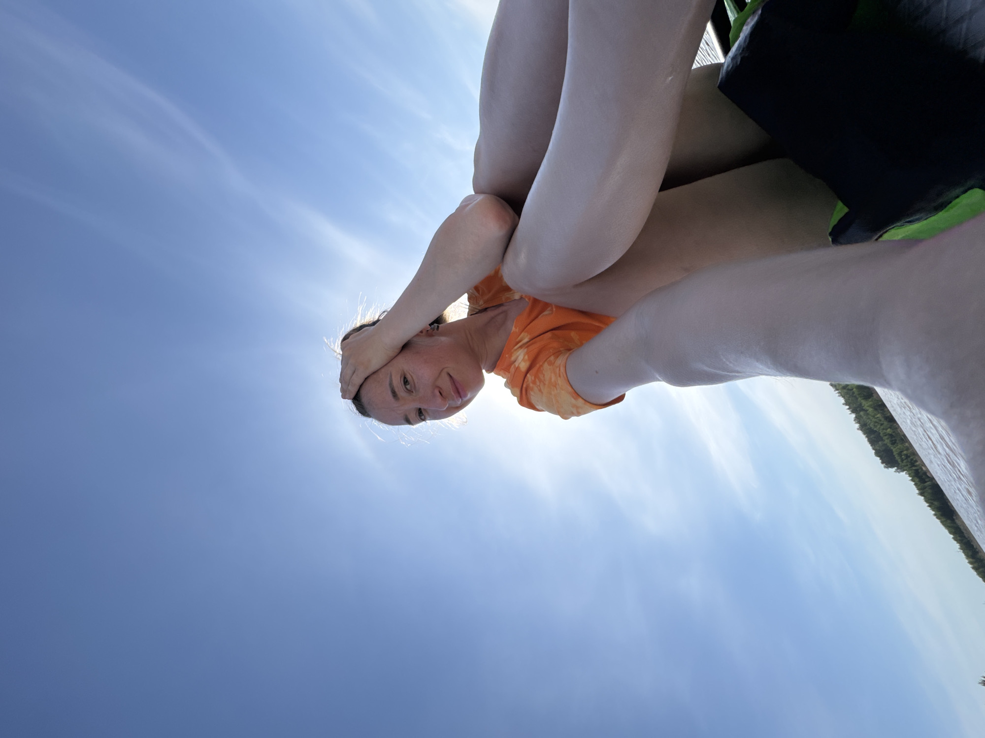 A woman with fair skin and brown hair is sitting on a black and green object. She is wearing an orange tie-dye t-shirt and has her hand on her forehead. The sky is blue with wispy clouds. In the background, there is a body of water with trees lining the shore. The image was taken from a low angle, looking up at the woman and the sky. Novyi, Russia is not visible in the picture.