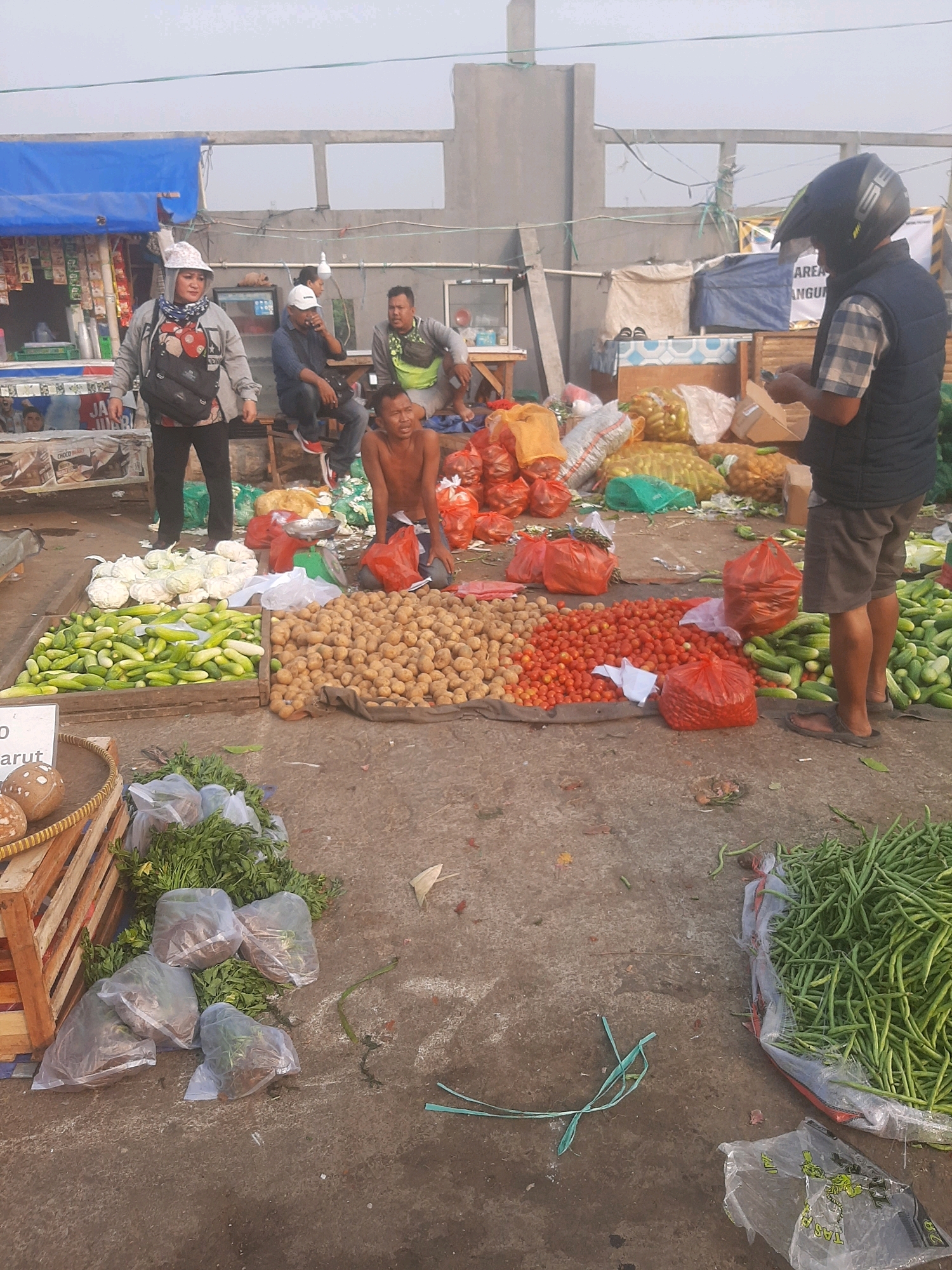 An outdoor market scene in Bekasi, Indonesia, displays a variety of fresh produce. In the foreground, a large pile of green beans is in a plastic bag, and to the left, a wooden crate holds cucumbers. Next to the cucumbers is a mound of potatoes and a pile of tomatoes. Cabbage heads are stacked on a tarp behind the cucumbers. A man in a vest and helmet stands to the right, facing away from the camera and looking at his phone. Behind him, a person is sitting on a bench. A shirtless man kneels in the center of the frame, looking towards the viewer. To the left, a woman wearing a hat and a scarf stands with her back to the camera, facing a market stall with blue tarpaulin. The ground is paved with concrete, and several plastic bags are scattered around. The background shows various market stalls and structures under a bright sky.