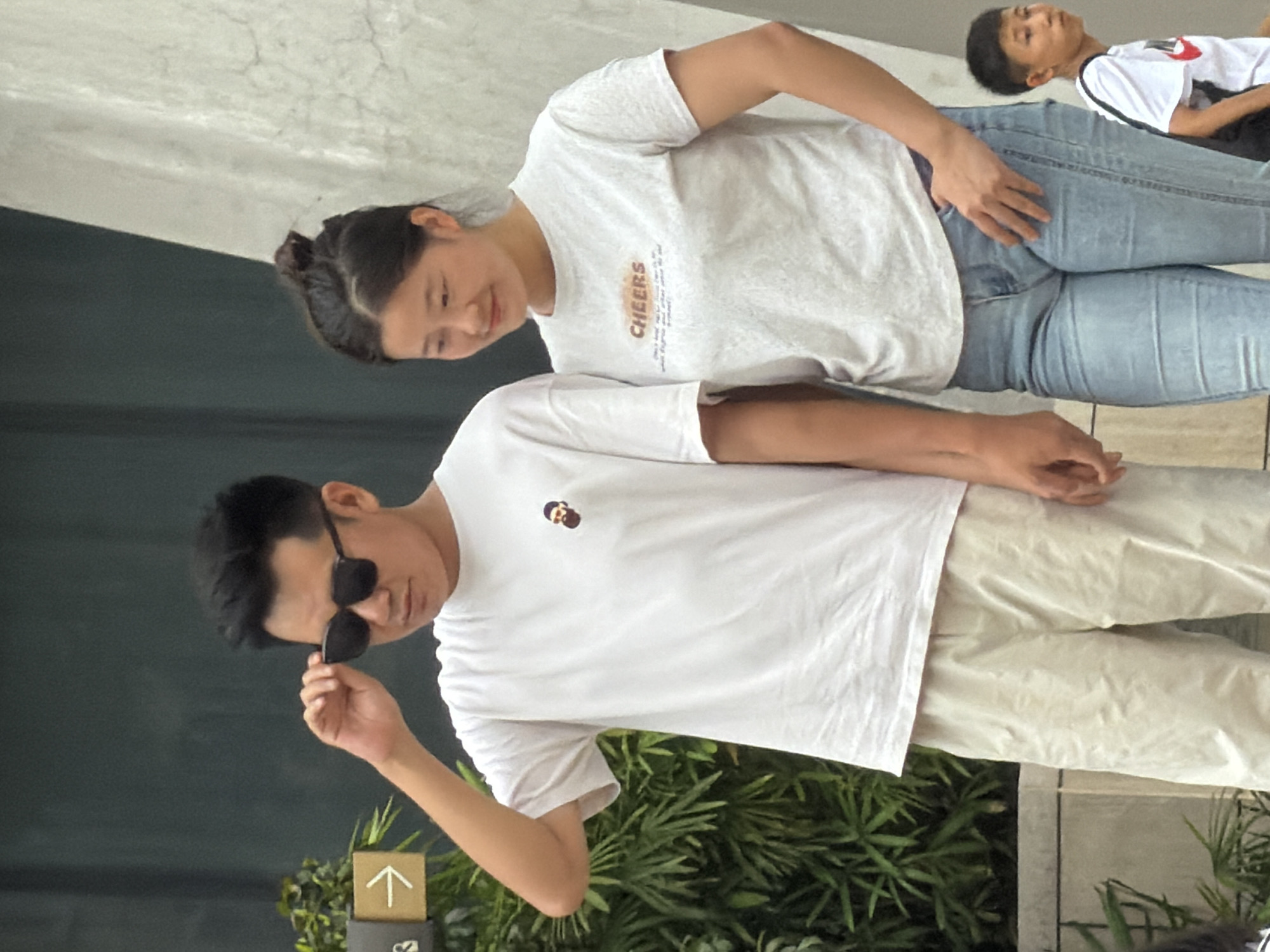 A medium shot captures a young Asian man wearing sunglasses and a white t-shirt with a small graphic of a man's head, standing to the left. He adjusts his sunglasses with his right hand. To his right stands a young Asian woman wearing a light grey t-shirt with the word "CHEERS" printed in orange and black letters, and light blue jeans. Her hair is pulled up in a bun. In the background, a young Asian boy in a white t-shirt with a red Nike logo is partially visible on the right. The scene is set outdoors, with lush green foliage and plants to the left and a textured concrete wall to the right. A directional arrow sign is also visible to the left of the man. The overall environment suggests an urban setting, possibly in Yuzhong District, China.