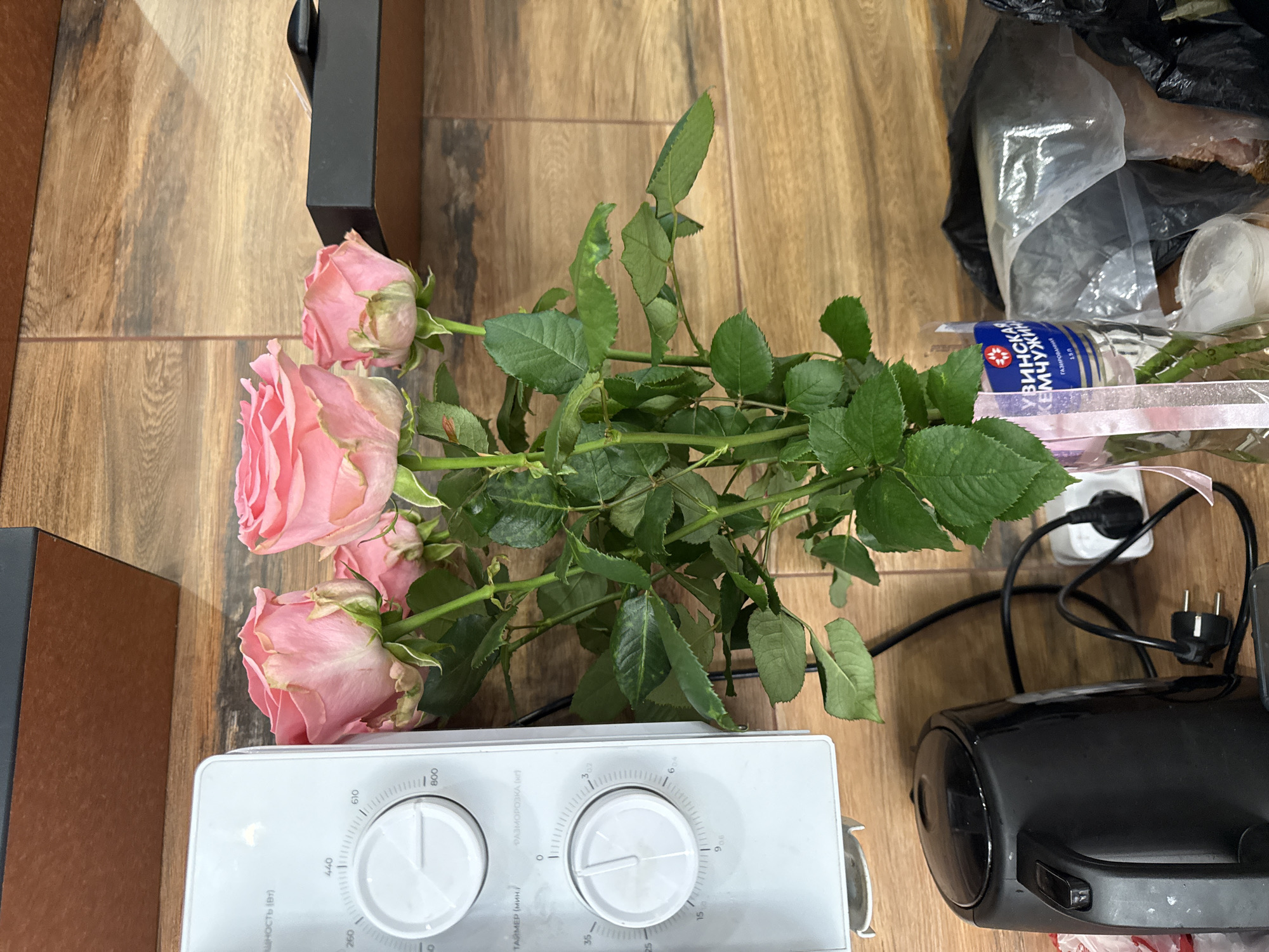 The image captures a still life scene with a bouquet of pink roses laid horizontally across a wooden surface, likely a floor or tabletop. The roses, in various stages of bloom, have vibrant green leaves, some showing minor signs of wear.
In the foreground, to the bottom left, a white kitchen appliance, possibly a microwave oven, is visible. It features two prominent circular dials. The left dial is marked with power settings in Russian: "МОЩНОСТЬ (Вт)" (POWER (W)) with values like 260, 440, 610, and 800. The right dial functions as a timer, labeled "ТАЙМЕР (МИН)" (TIMER (MIN)), with settings up to 35 minutes, and also indicates "РАЗМОРОЗКА (кг)" (DEFROST (kg)) with weight options up to 3.02 kg.
To the bottom right, a black electrical appliance, potentially a handheld vacuum cleaner or similar device, is present, accompanied by black power cords and a plug. In the upper right corner, a transparent plastic bag containing various items, including a bottle, is seen. The bottle has a blue label with Cyrillic text, which reads "УВИНСКАЯ КЕМЧУЖИНА ГАЗИРОВАННАЯ 1.5Л," indicating it is 1.5 liters of "Uvinskaya Kemchuzhina" carbonated water. A pink ribbon is also visible near the bottle and flowers. Part of a black frame or object is partially visible on the top left. The presence of Cyrillic text on the appliance and bottle suggests the location is in Russia, consistent with the provided context of Chaikovsky.