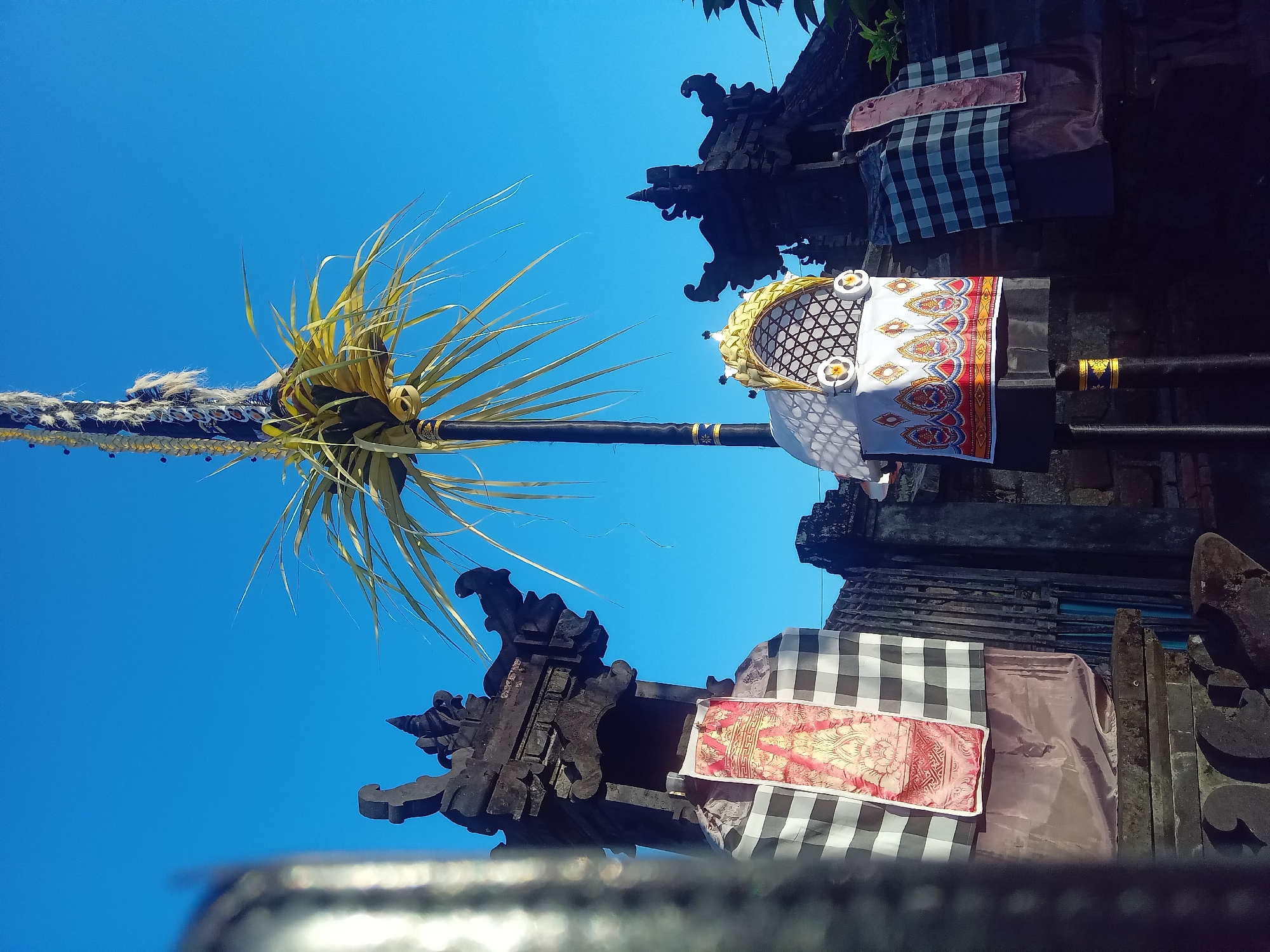 In Blahbatuh, Indonesia, on a clear blue sky day, a decorative bamboo pole stands tall, adorned with woven palm leaves at its crown. Below, two stone temple gates are draped with traditional black and white checkered cloth, known as "kamben gilit." One gate features a vibrant red and gold textile depicting intricate patterns. Another, more ornate structure, also decorated with the checkered cloth, is positioned in front of the stone gateway. This scene captures a glimpse of cultural and religious practices in Bali, likely during a festival or ceremony.