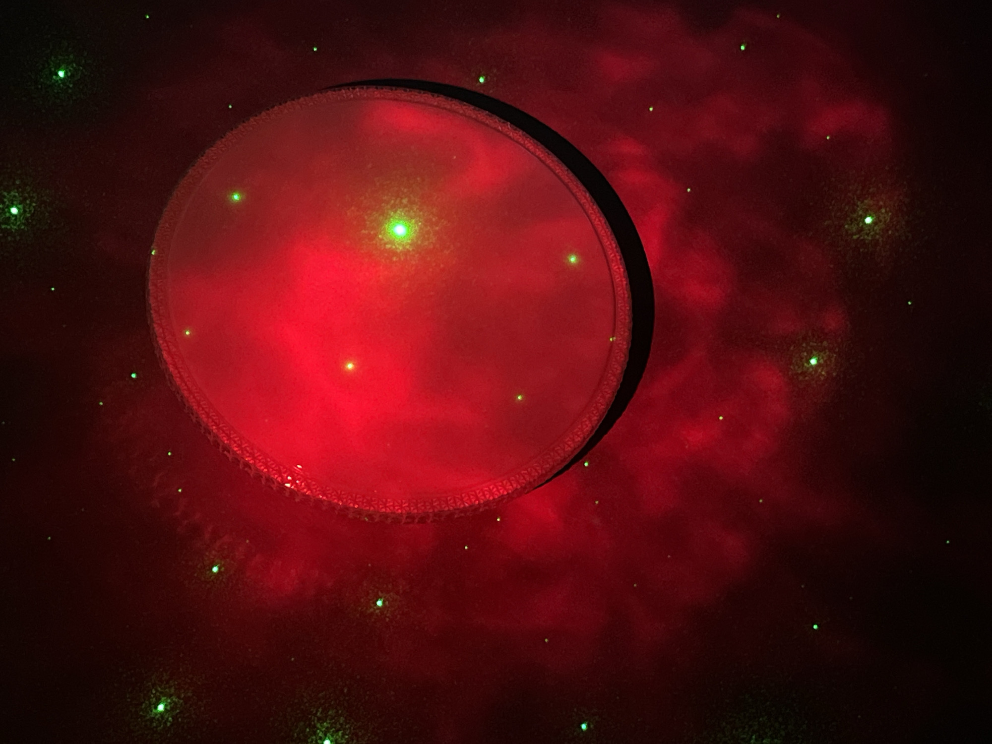 The image shows a dark room illuminated by a red light source, possibly a projector, casting a circular shape onto a wall or surface. The circle is surrounded by a dark outline, suggesting a physical object projecting the light. Scattered green laser dots are visible across the red light and surrounding darker areas, creating a star-like effect. The background has a blurred, nebula-like appearance with varying intensities of red. The room appears to be indoors, and the overall atmosphere is dim and otherworldly. There is no information in the image that indicates the location is Nizhny Tagil, Russia, or the involvement of people or specific actions.