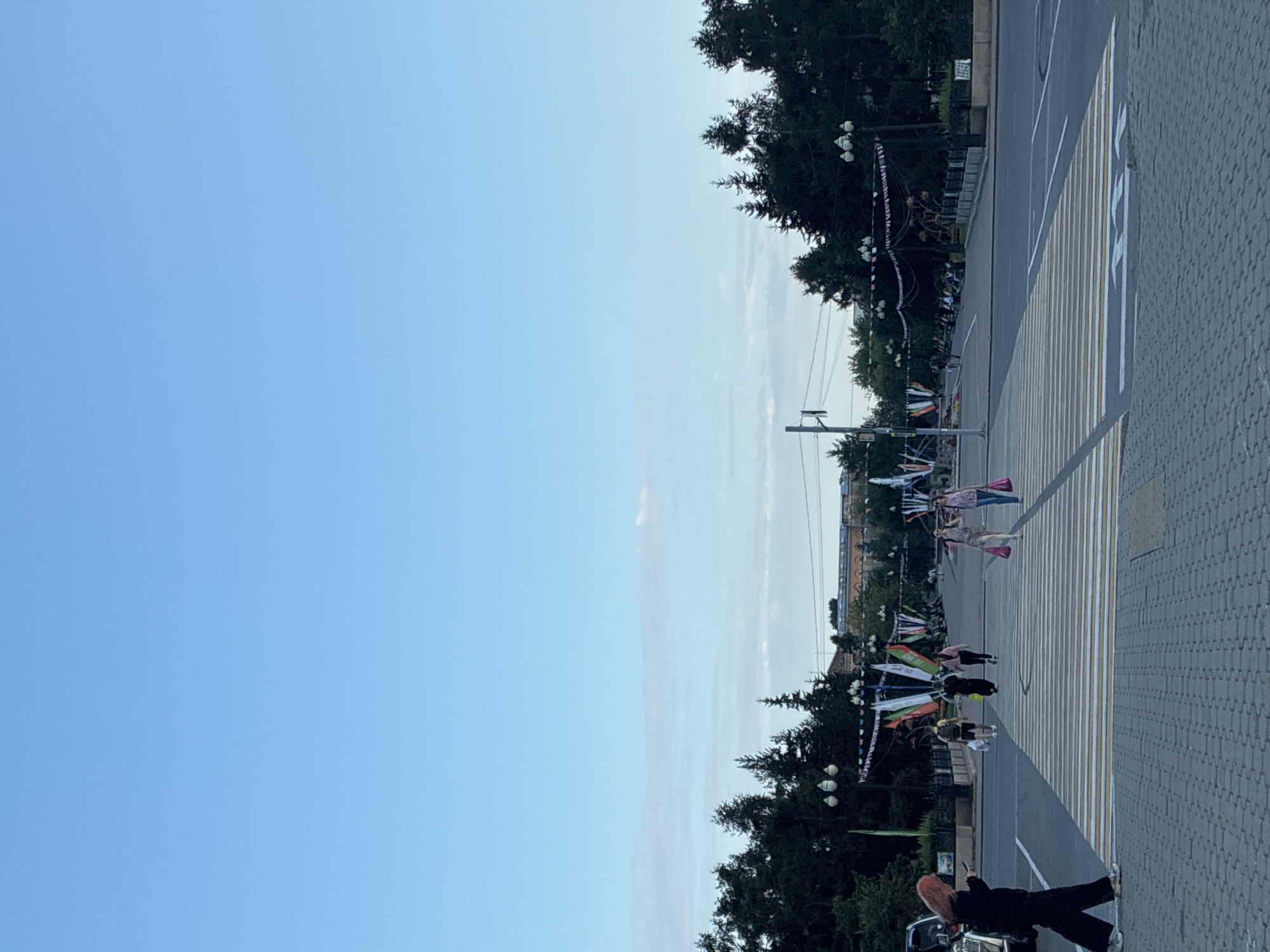 This is an eye-level, medium shot of a wide pedestrian crossing in a city, possibly Irkutsk, Russia, given the context. The sky is a clear, pale blue with a few wispy clouds near the horizon. A double row of trees lines both sides of a wide avenue in the background, with flags and bunting strung between lampposts. In the far distance, a large, light-colored building with arched windows is visible.
On the pedestrian crossing in the foreground, several people are present. A person with bright orange hair, wearing black clothing, is walking across the street from left to right, looking down at their phone. Further across the crossing, two individuals are walking together, one carrying two pink shopping bags. Behind them, several other people are crossing or walking on the avenue. A few parked cars are visible on the left side of the image. The ground is paved with interlocking grey bricks.