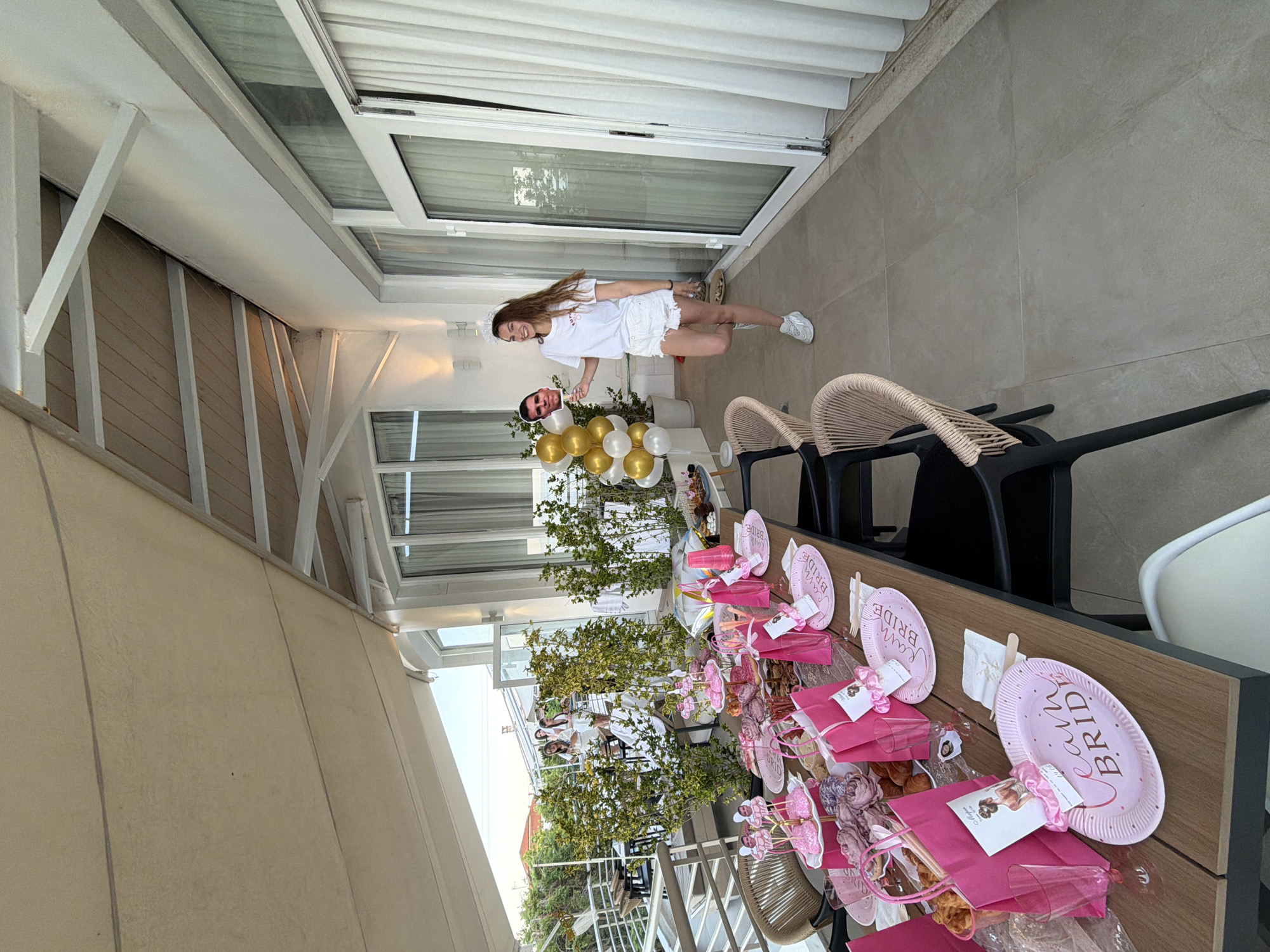 A young woman with long brown hair, wearing a white t-shirt and white shorts, stands on a patio in Kassandra Municipal Unit, Greece. She is holding a cutout of a man's face on a stick and a tiara on her head, suggesting a bachelorette party. Behind her, a table is laden with pink party decorations, including plates, gift bags, cups, and balloons. The patio overlooks a balcony with lush green plants and other guests in the background. The setting appears to be a bright, sunny day.