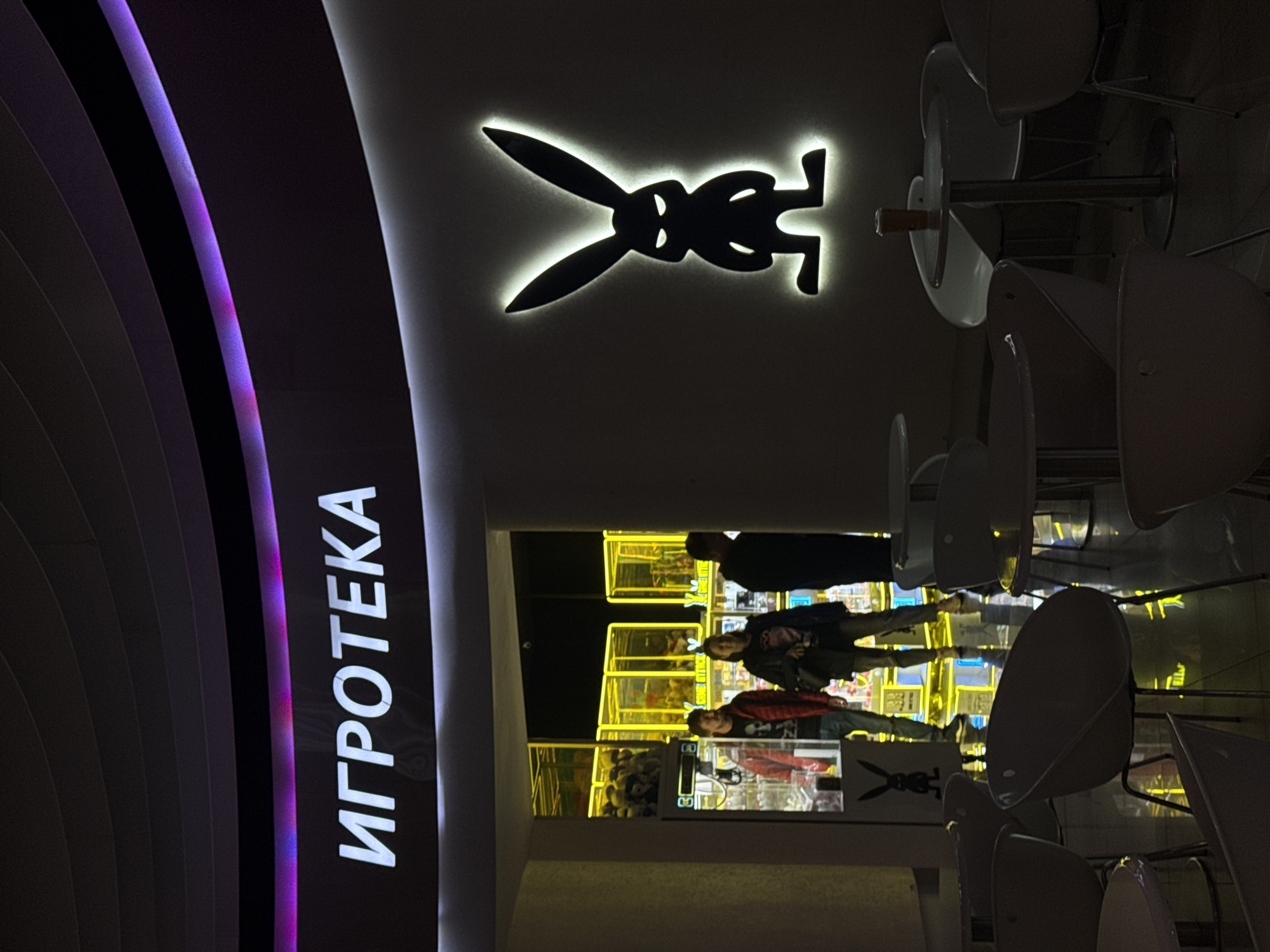 The image depicts the entrance of a store in Moscow, Russia, called "ИГРОТЕКА," which translates to "Game Room" or "Arcade." A stylized, illuminated black logo featuring a bunny with legs extends from the wall. Inside the store, three individuals stand near claw machines. Chairs with white seats are positioned outside the storefront. The lighting is subdued with neon accents on the storefront.