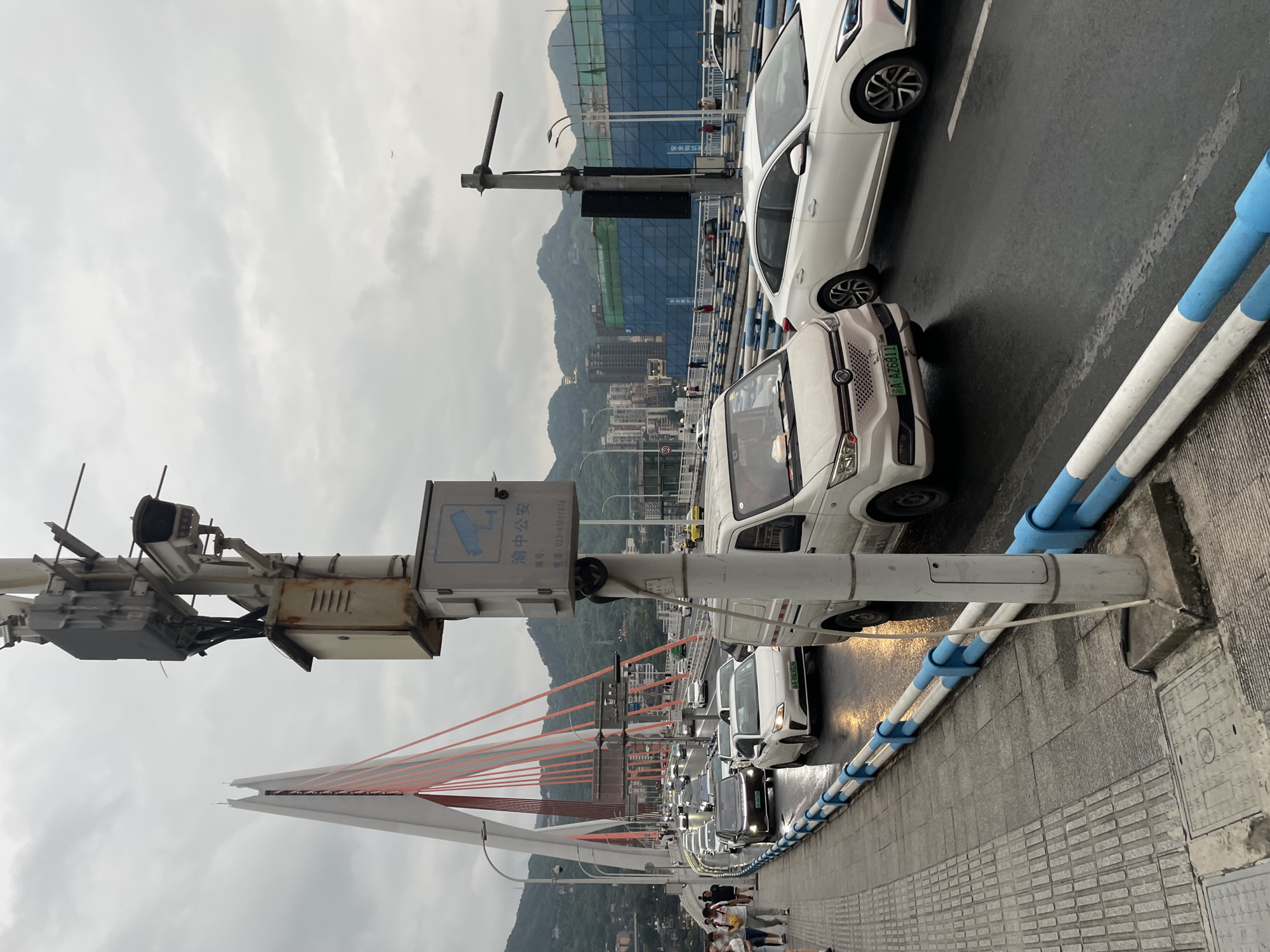 The image depicts a view of the Hongyadong Bridge in Yuzhong District, China, during overcast weather. The bridge features prominent white towers and orange suspension cables. A busy roadway crosses the bridge, filled with a line of vehicles, including cars and a white van. The road surface appears wet, suggesting recent rain. Pedestrians are visible on the sidewalk to the left of the road. On the right side of the image, a tall pole with a surveillance camera and a box containing electronics is mounted near the edge of the bridge. The background shows a cityscape with buildings nestled among lush, green hills, and construction sites covered in blue netting. The overall scene conveys a sense of urban activity and infrastructure within a natural landscape.