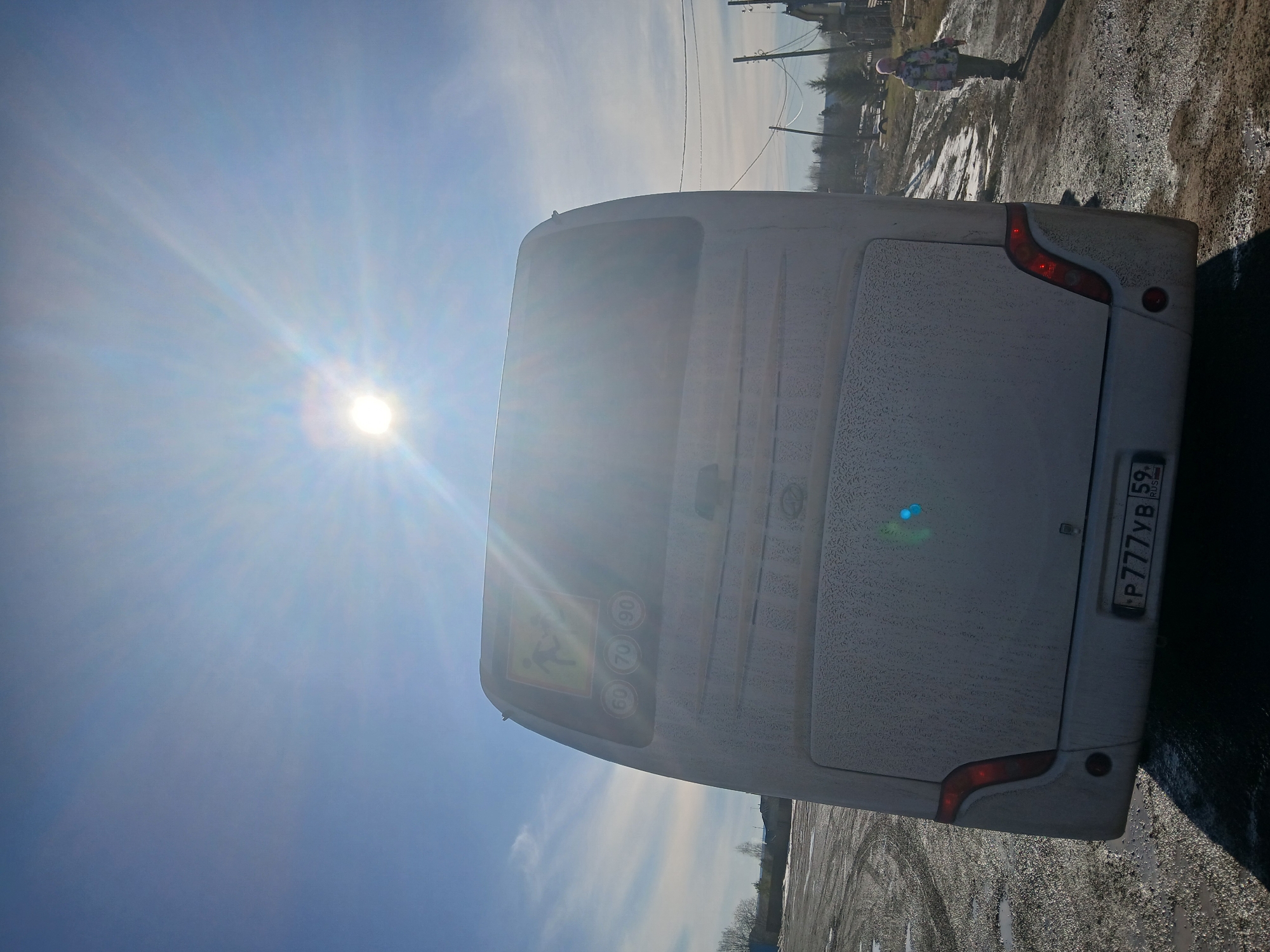 A white bus, bearing the license plate number P777YB 59, is the primary subject, positioned on a surface that appears to be asphalt or a similar material. The surface is mottled, with indications of tire tracks and variable coloration. The bus is partially covered with a granular substance, suggesting possible frost or similar environmental deposits.
The sky is bright blue with a strong, direct sunlight source causing significant lens flare. In the background, there are structures that seem to be buildings or houses, with power lines visible above them. A figure stands distantly on the side of the road.
The location is in the city of Unknown, Unknown.