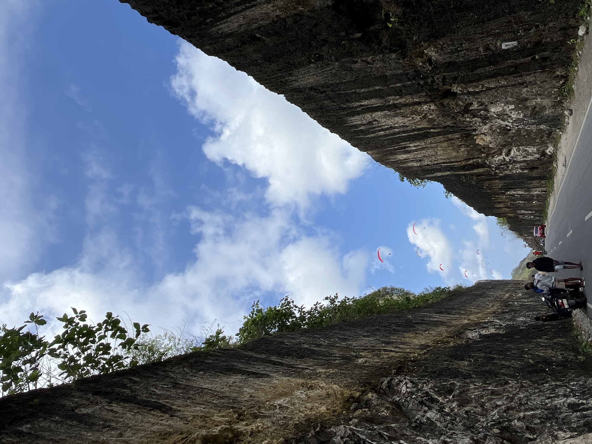 The image captures a scenic view of a road cutting through two towering, rugged cliffs. The cliffs are textured with shades of gray and brown, with sparse vegetation growing from their surfaces. Above, a vibrant blue sky is dotted with fluffy white clouds, and several paragliders are visible, appearing as small colorful dots against the expansive sky. In the foreground, a paved road runs between the cliffs. On the side of the road, several people are gathered around a motorcycle. A red car is also visible further down the road. The scene suggests a popular tourist destination in Kuta Selatan, Indonesia, likely known for its paragliding activities and dramatic natural landscapes.