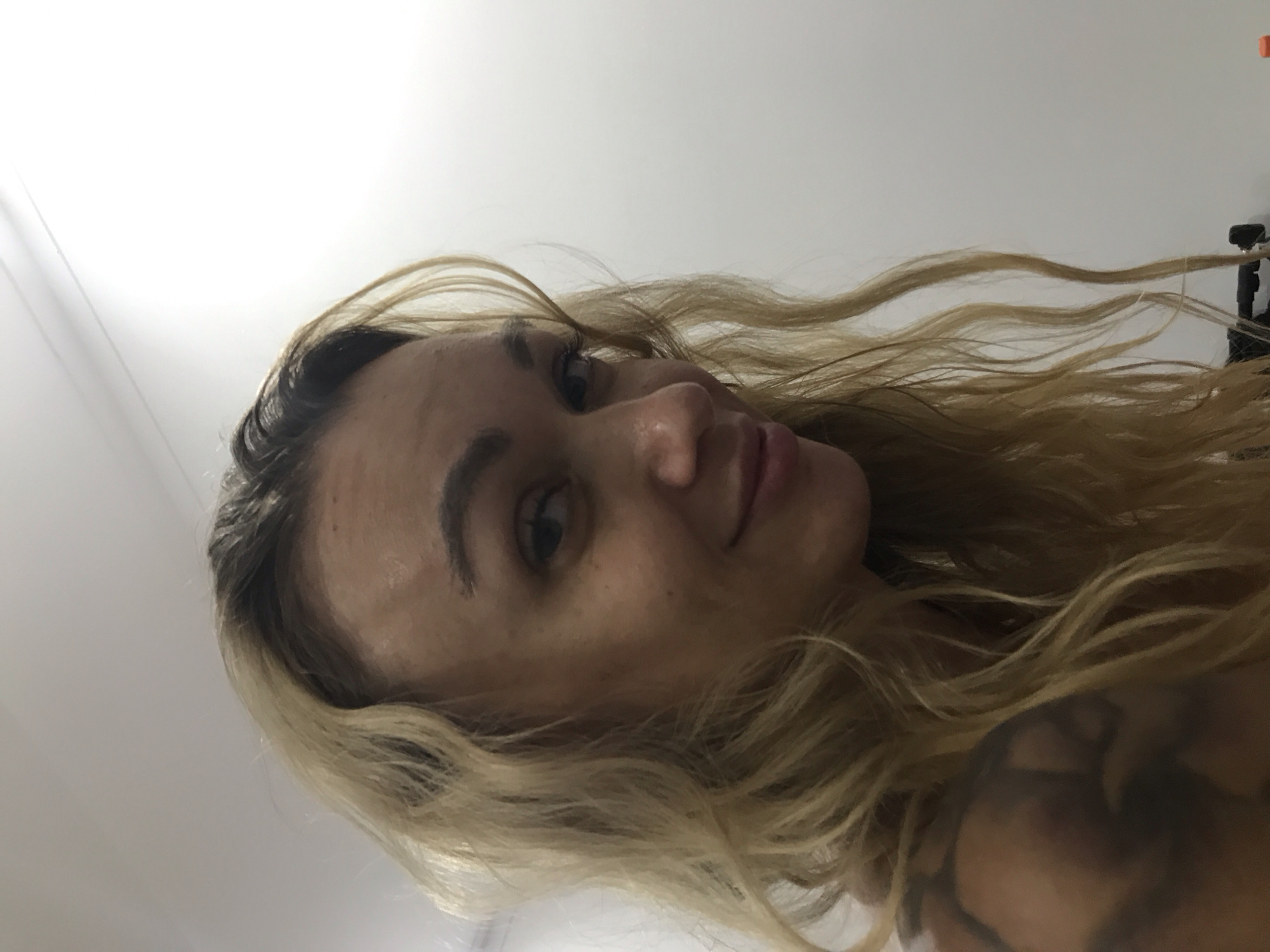 This close-up, low-angle portrait features an adult woman with long, wavy blonde hair and visible darker roots. The image is rotated 90 degrees counter-clockwise. She is looking towards the camera with a subtle smile or smirk, her right eye (viewer's left), nose, and lips clearly visible. Her skin has a natural appearance with some freckles or minor blemishes. A dark-ink tattoo is partially visible on her shoulder or upper arm, obscured by her flowing hair. In the upper right portion of the frame, amidst her hair, a dark object resembling a hair clip or a small camera accessory can be seen against a plain, light-colored background, likely an indoor wall or ceiling. The lighting is bright and even, consistent with an indoor setting. The woman's expression conveys a calm or mildly amused demeanor, captured in what appears to be a casual selfie, potentially taken in Mũi Né, Vietnam.