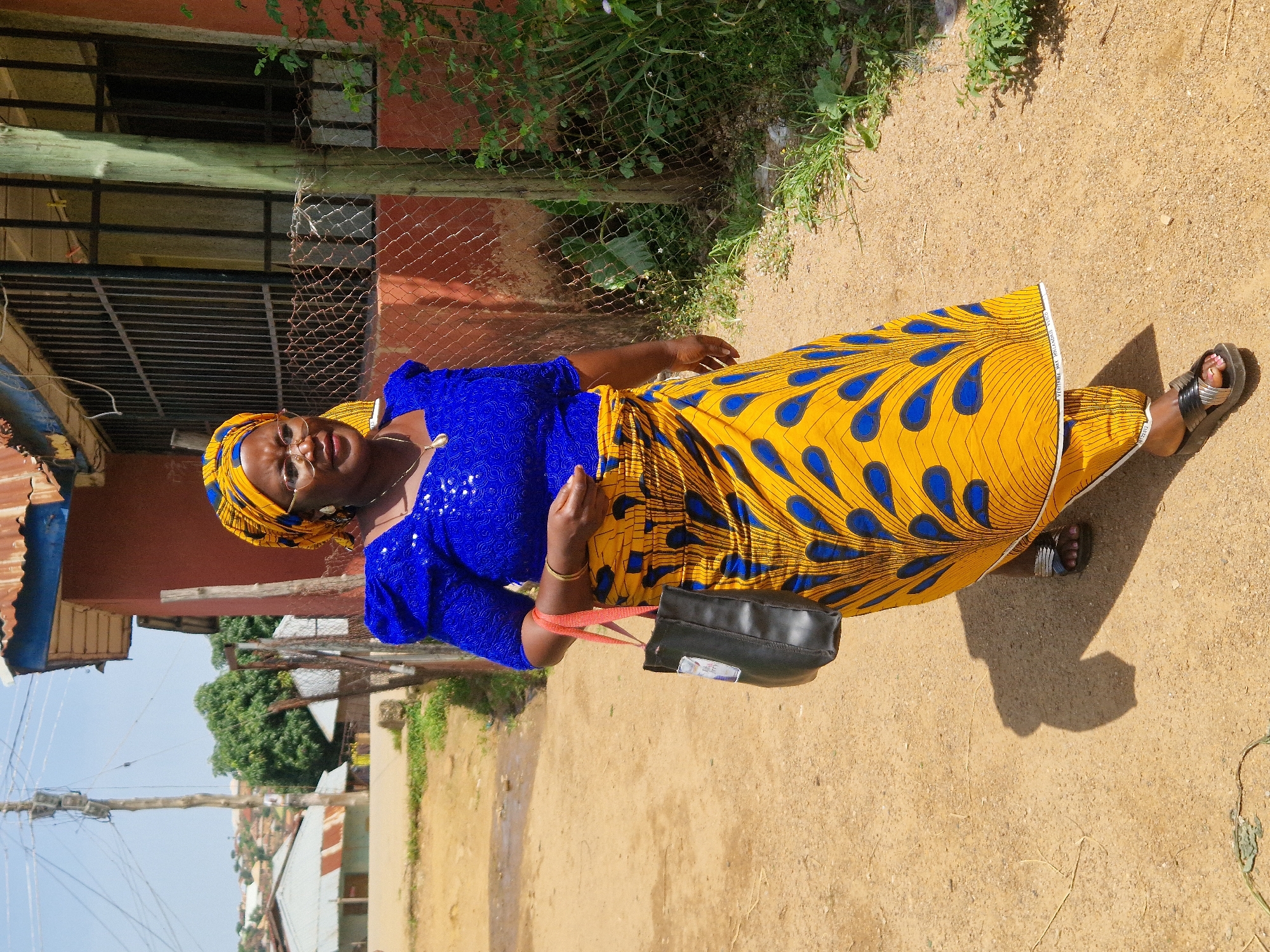 A medium shot shows a Black woman walking towards the camera on a dirt path in an outdoor, urban environment. She is wearing a bright blue, sequined, short-sleeved top and a yellow wrap skirt with a blue peacock feather pattern. A yellow and blue patterned headwrap covers her hair. She has on glasses and gold earrings. Around her neck, she wears a gold chain with a small gold pendant. She is carrying a black tote bag with red straps over her left shoulder. On her feet, she wears black slide sandals. The background shows a building with a red facade and a barred window, a chain-link fence, and some greenery. Further in the background, there are other buildings and houses on a hillside under a clear blue sky. The lighting suggests it is daytime.