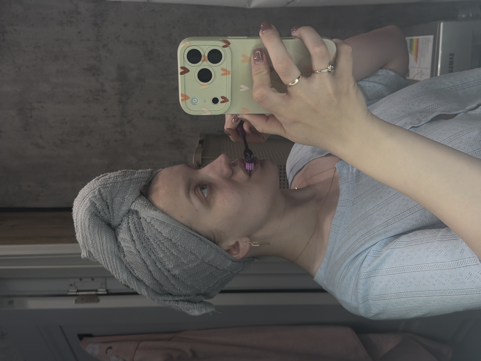A woman is brushing her teeth while taking a selfie in what appears to be a bathroom. She is wearing a grey towel wrapped around her head and a light blue, patterned shirt. The phone she is holding is light green with three camera lenses. Her hand has two rings on it. The background is blurry, but it appears to be a wall and a cabinet. The location in the city of Reshetnikovo, Russia, is not identifiable.