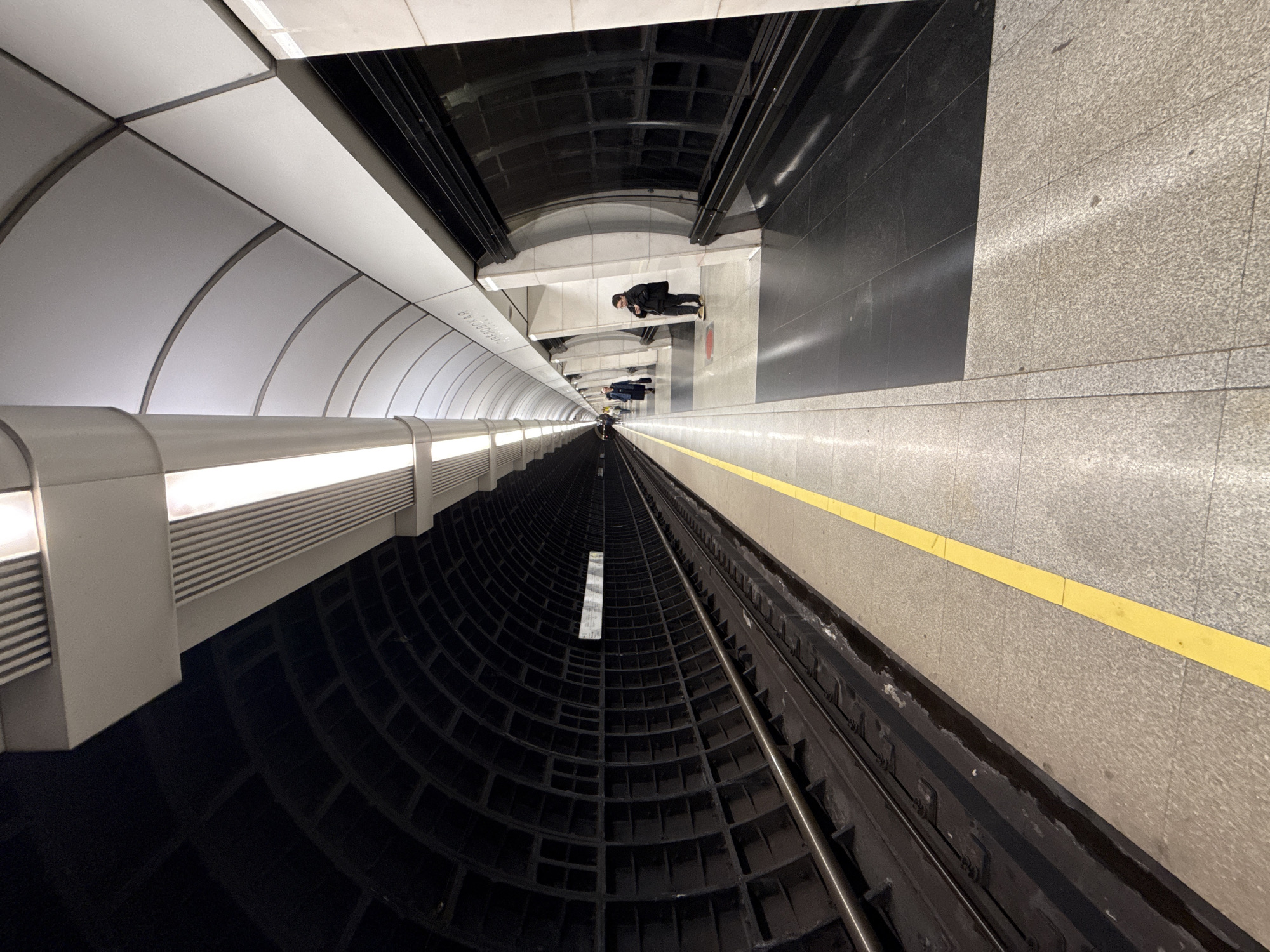 This image captures a disorienting perspective of a Moscow metro station. The viewpoint is oriented at an unusual angle, looking down the platform towards the tunnel. People are present on the platform, standing and looking at their phones. The platform features gray tiled floors and a yellow safety line. The opposite wall is finished with black and white tiles. Between the platform and the wall are the subway tracks. The tunnel is black and appears to be constructed of curved, gridded concrete.