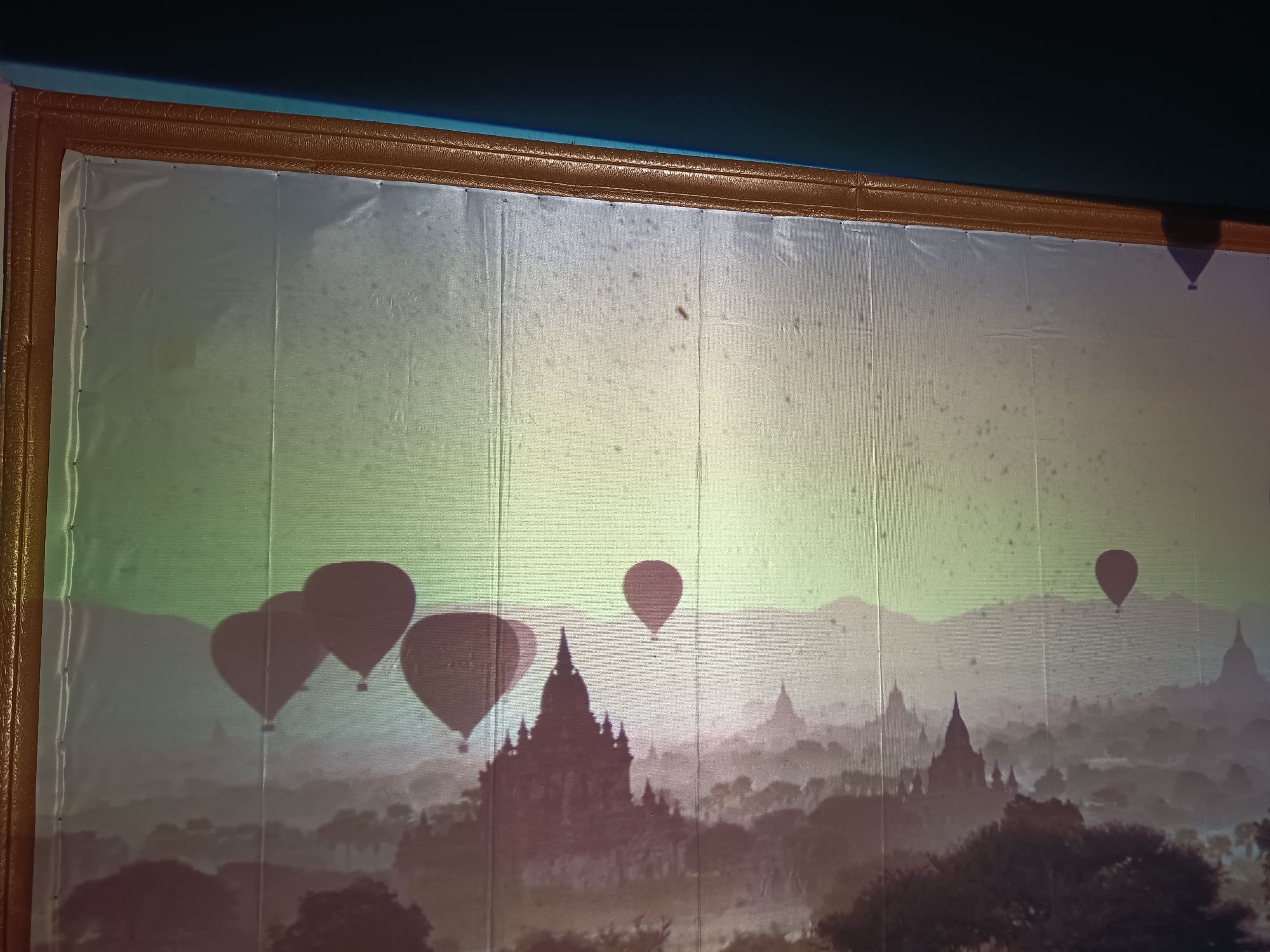 The image displays a framed panorama of a misty landscape, possibly a digital projection or a large print. The scene depicts numerous hot air balloons floating above a silhouetted city or temple complex with distinctive pagoda-like structures. The balloons are various shades of dark red and purple, some appearing as solid silhouettes, others slightly translucent. The landscape itself is rendered in muted tones of green, gray, and brown, with rolling hills or mountains in the background obscured by fog. The foreground is filled with dark silhouettes of trees. The overall impression is one of an atmospheric dawn or dusk. The location in Kalipenggung, Indonesia is not discernible from the visual content of the image.